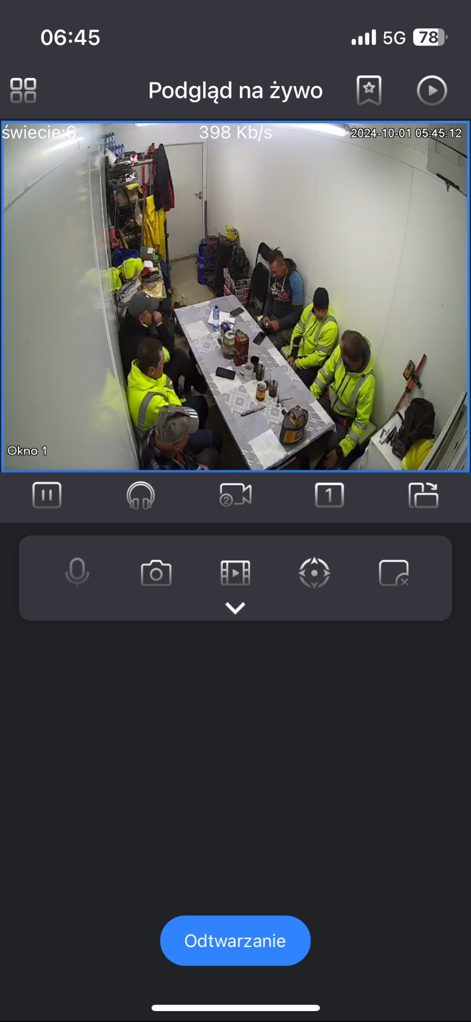 Podgląd z kamery monitoringu: grupa osób w odblaskowych kamizelkach siedzi przy stole w pomieszczeniu. Widok z aplikacji mobilnej, data i godzina w prawym górnym rogu.