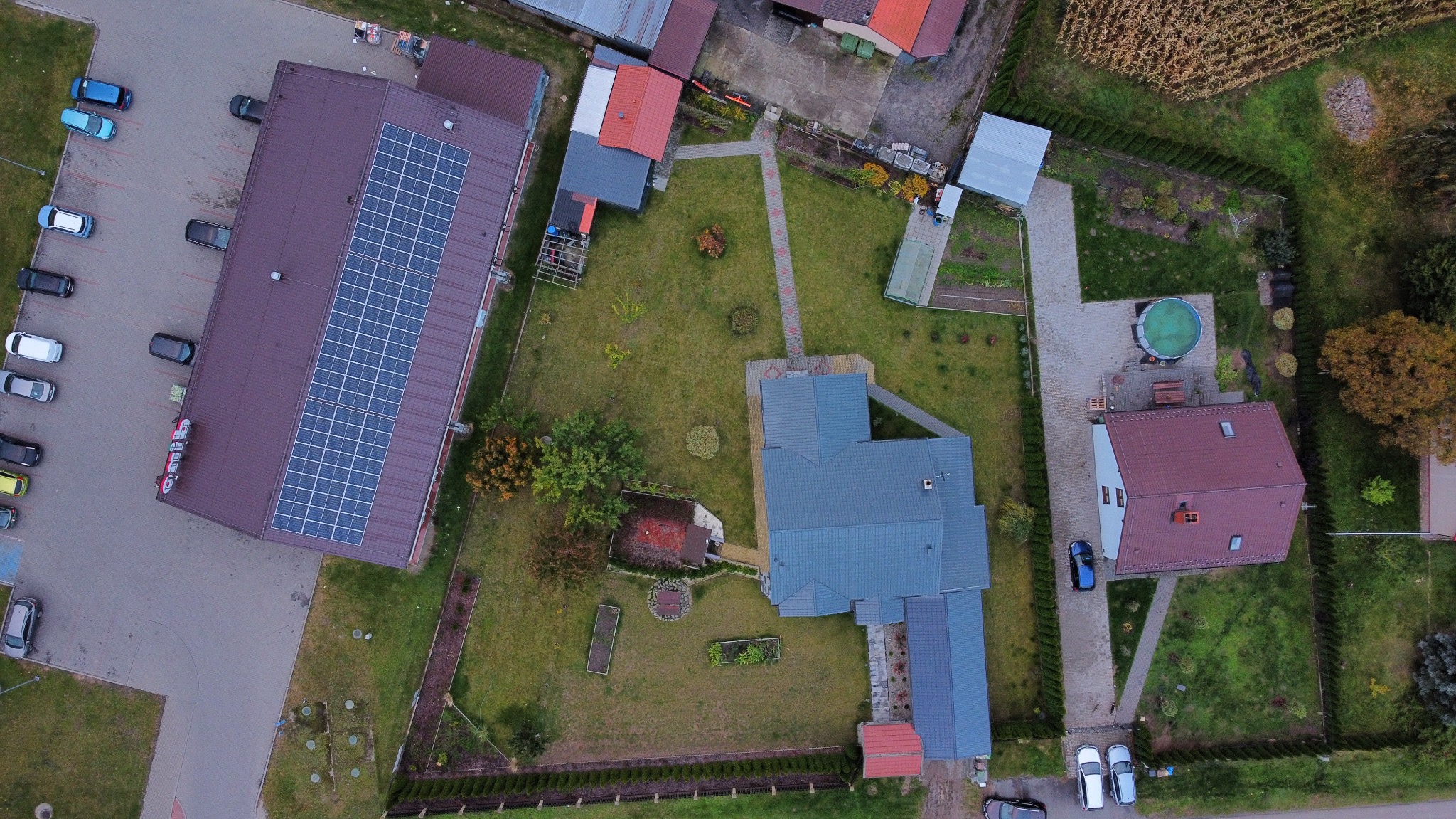 Widok z drona na budynek z panelami słonecznymi na dachu, otoczony zielenią i innymi zabudowaniami. Ujęcie z góry ukazuje całą posesję i jej otoczenie.