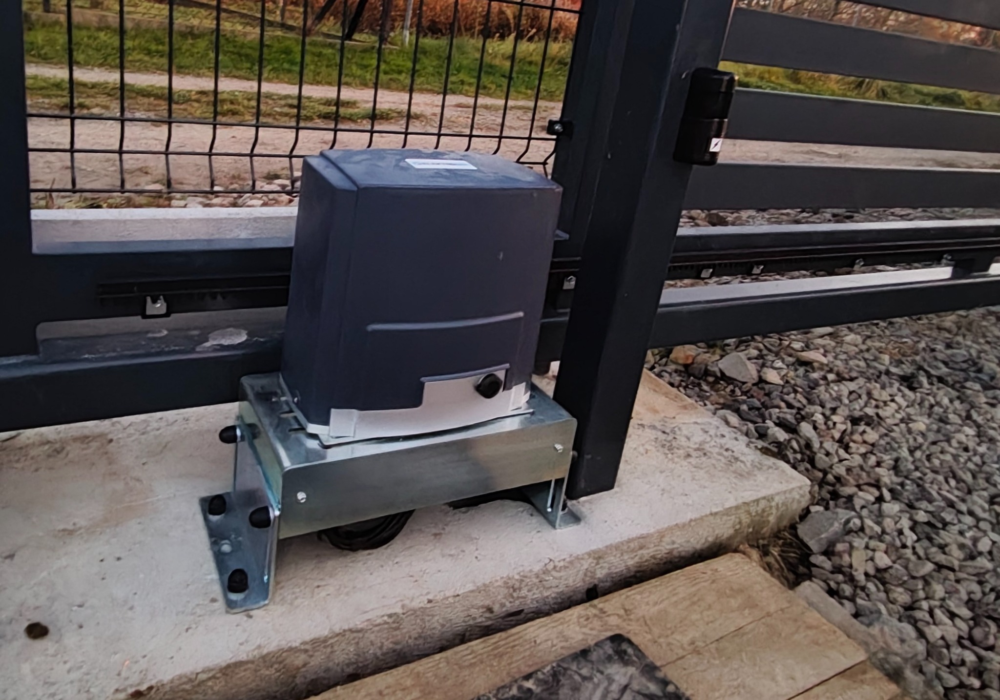 Szary automat do bramy przesuwnej, zamontowany na betonowej podstawie, z widocznymi elementami mocującymi i fragmentem ogrodzenia w tle.