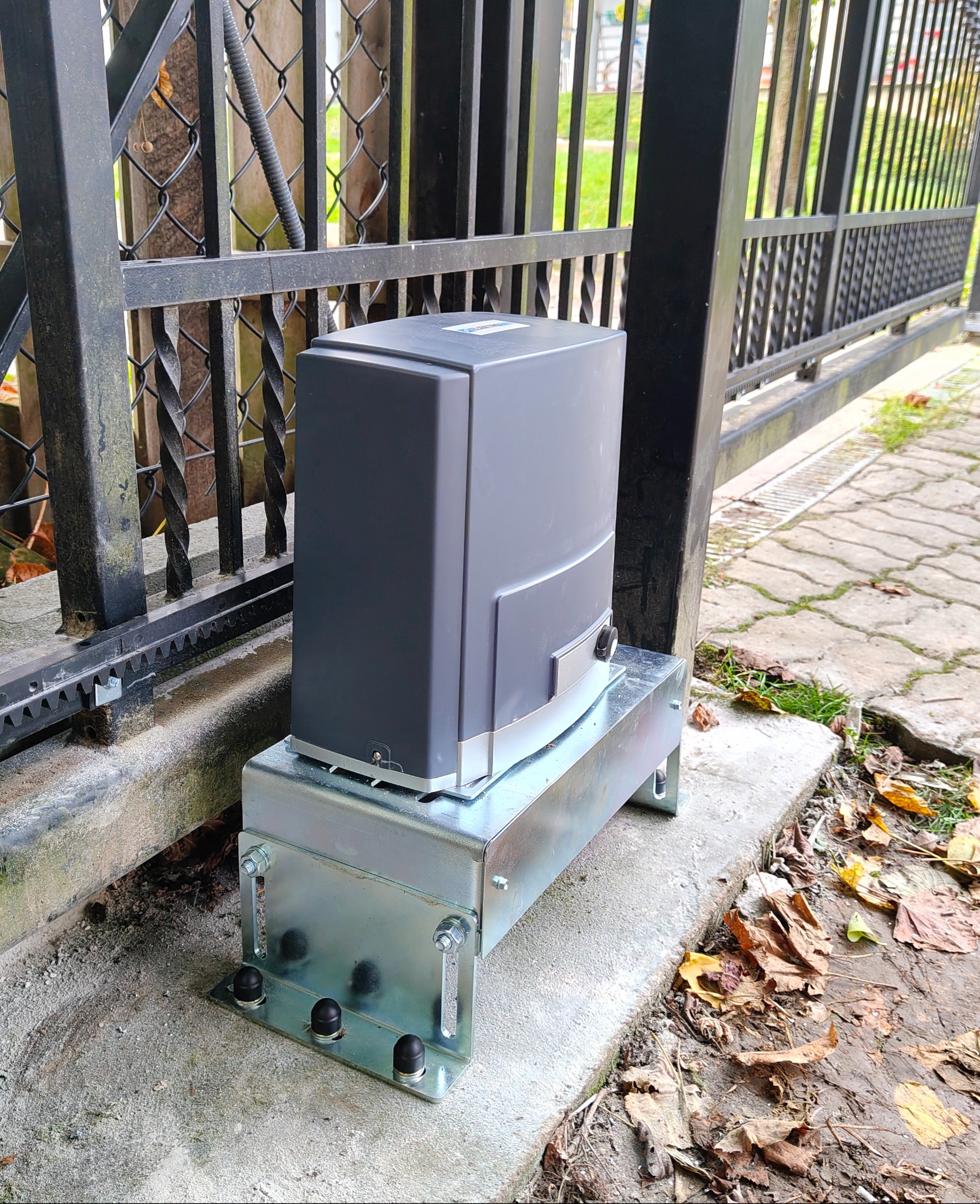 Szary napęd do bramy przesuwnej zamontowany na metalowej podstawie w Bratkowicach. Widoczna zębatka bramy i kostka brukowa.