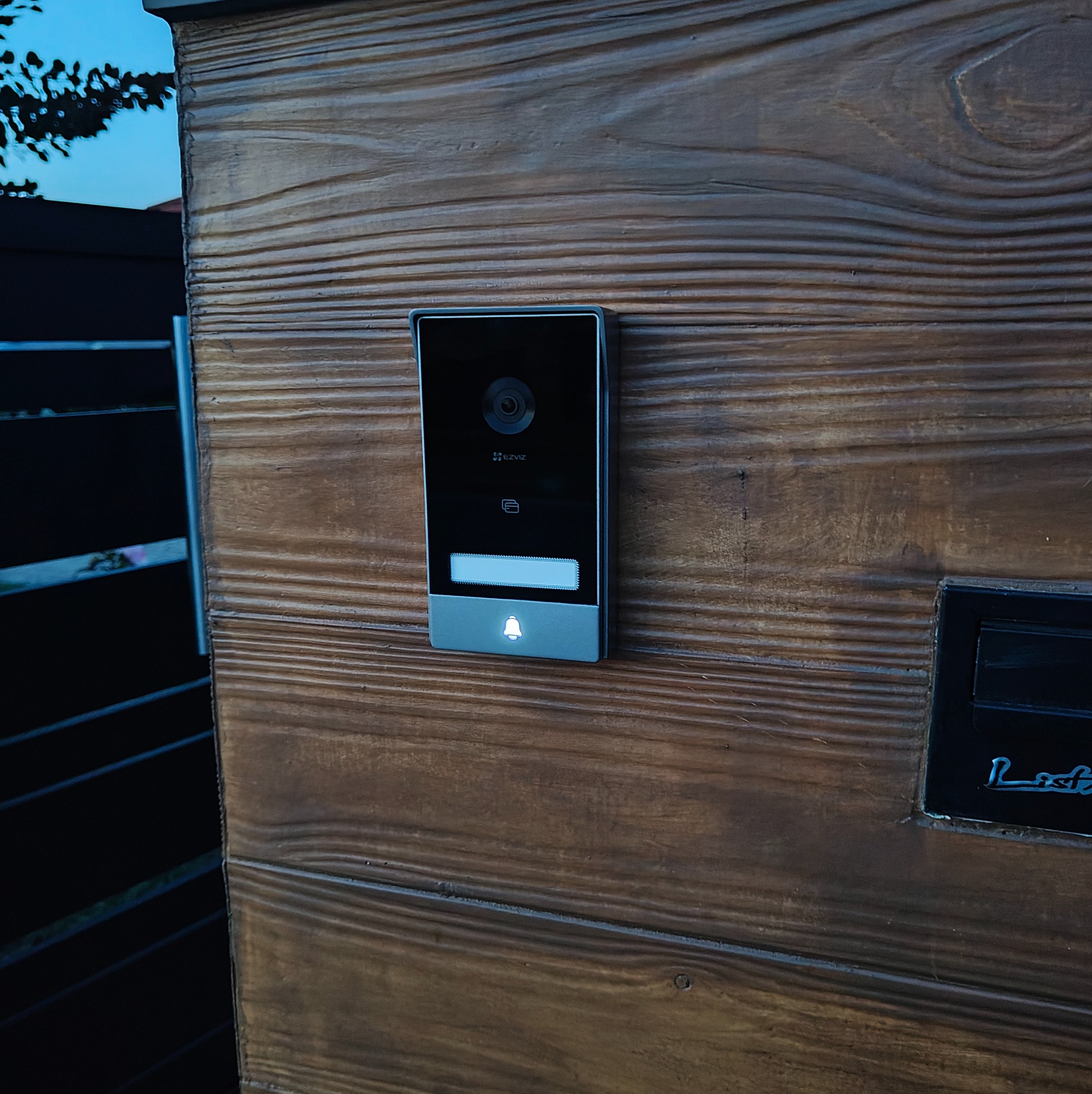 Nowoczesny wideodomofon EZVIZ zamontowany na drewnianej bramie wjazdowej z widocznym oświetleniem dzwonka i kamerą. Minimalistyczny design.