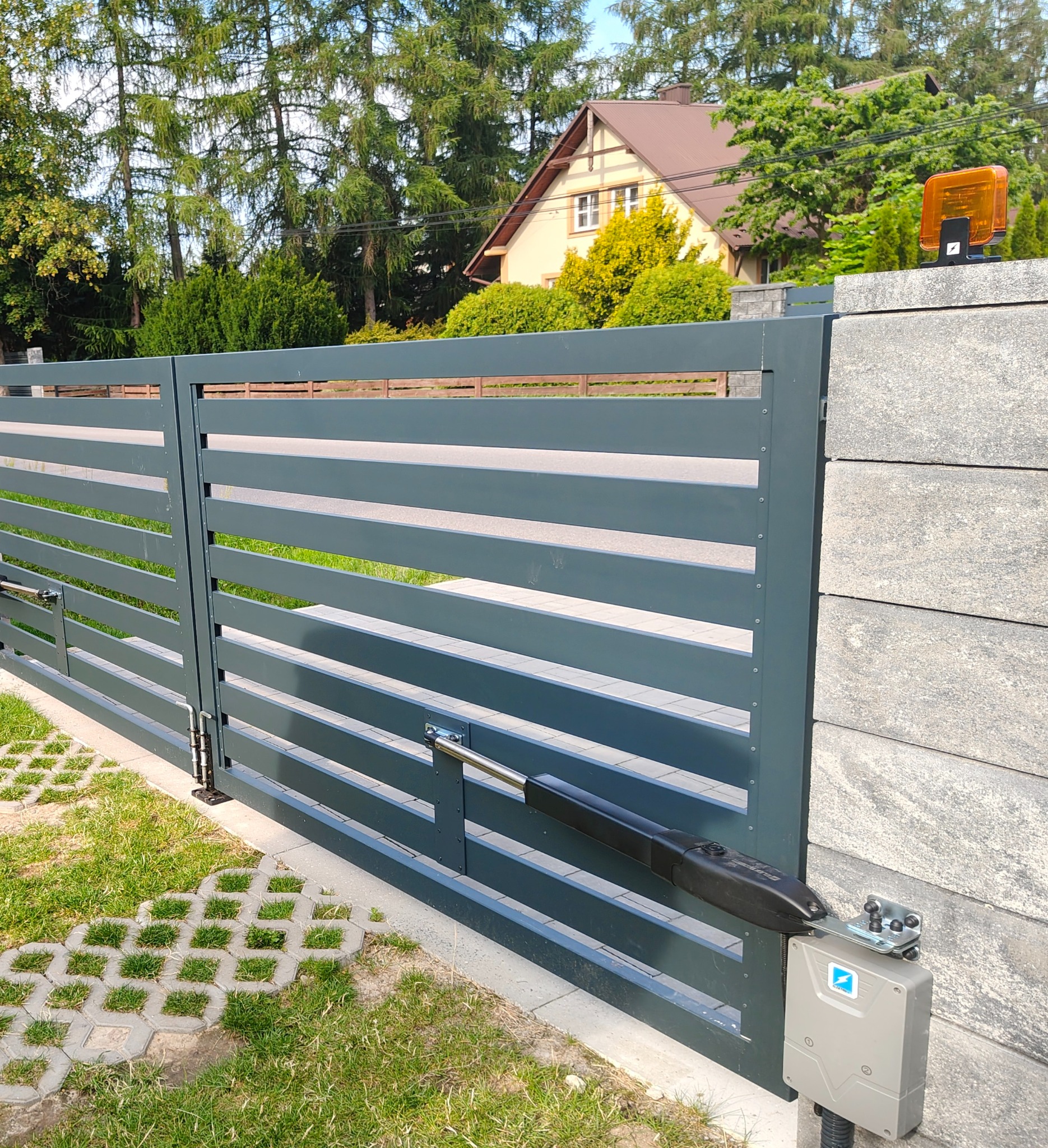Nowoczesna, pozioma brama wjazdowa z automatyką, szara, z widocznym siłownikiem i lampą ostrzegawczą. Trawa i kostka ażurowa przed bramą, dom w tle.