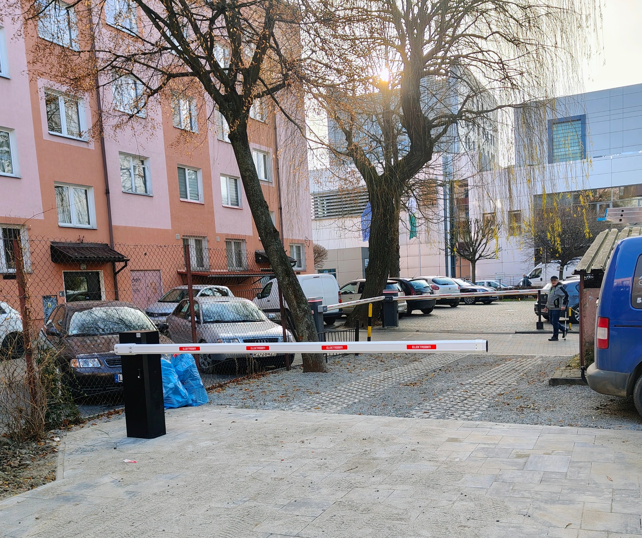 Szlaban automatyczny z napisem ELEKTROBIM na tle parkingu, budynków mieszkalnych i drzew. Widoczny fragment niebieskiego samochodu dostawczego.