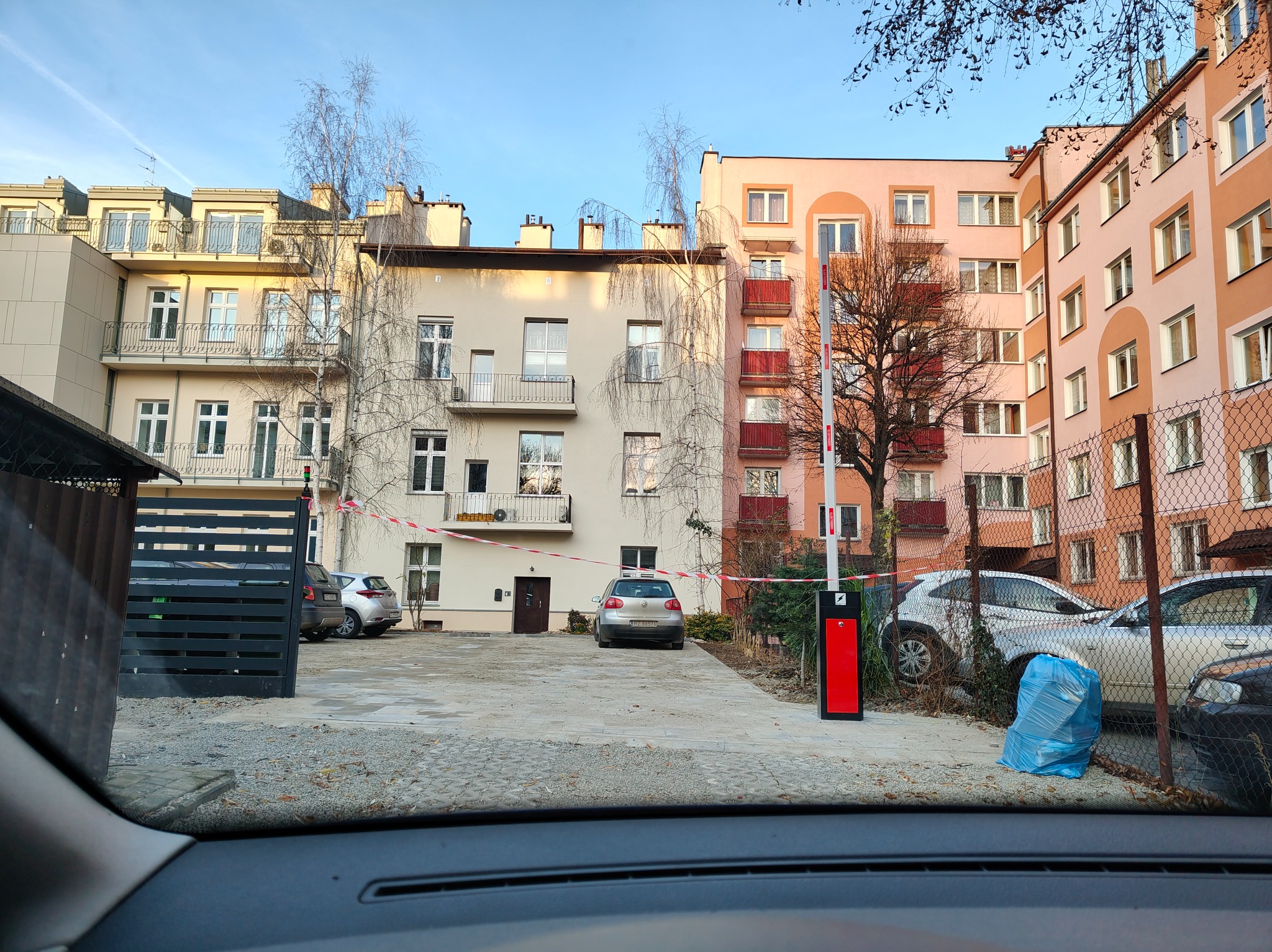 Szlaban automatyczny na wjeździe do osiedla mieszkaniowego, widok z wnętrza samochodu. W tle budynki mieszkalne i zaparkowane auta.