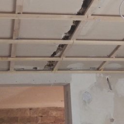 WILDBUD PAWEŁ HANAJ - Uszkodzony sufit podwieszany z widoczną konstrukcją drewnianą i fragmentem ściany z otworem drzwiowym w trakcie prac remontowych. Widoczne niedoskonałości i brak wykończenia.