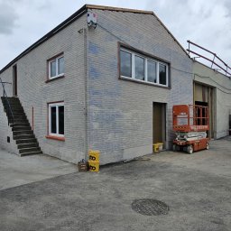 WILDBUD PAWEŁ HANAJ - Budynek z cegły w trakcie malowania elewacji na szaro, widoczne ślady po malowaniu, podnośnik koszowy i wiaderka z farbą. Częściowo pomalowana ściana.