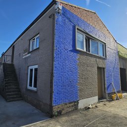 WILDBUD PAWEŁ HANAJ - Budynek z fragmentami elewacji w różnych kolorach (szary, niebieski, zielony), ukazujący postęp prac malarskich. Widoczne okna, schody zewnętrzne i sprzęt malarski.