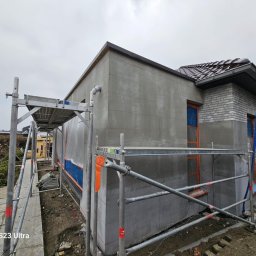 WILDBUD PAWEŁ HANAJ - Ocieplanie elewacji domu jednorodzinnego w Urzędowie. Widoczne rusztowania i przygotowana powierzchnia pod tynk. Część budynku wykończona cegłą.
