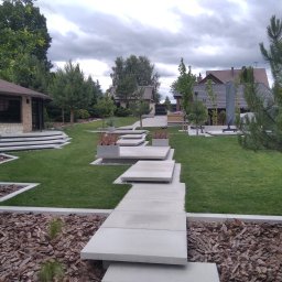 WILDBUD PAWEŁ HANAJ - Nowoczesny ogród z trawnikiem, ścieżką z płyt betonowych i rabatami z korą. W tle budynek z kamienia i cegły oraz domy jednorodzinne pod pochmurnym niebem.