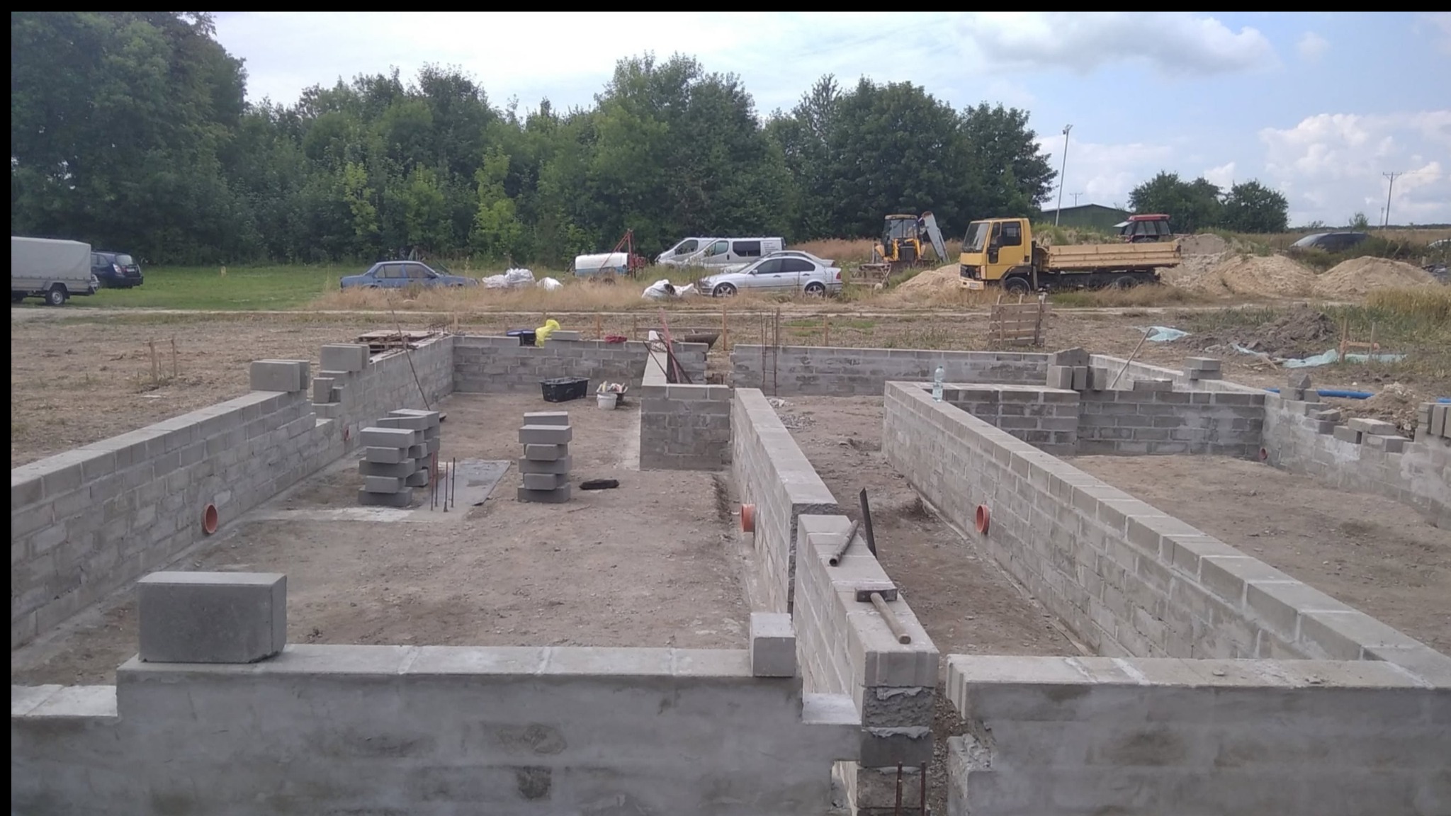 Fundamenty domu z bloczków betonowych na działce budowlanej, widoczne zbrojenia i otwory wentylacyjne. W tle sprzęt budowlany i zieleń.