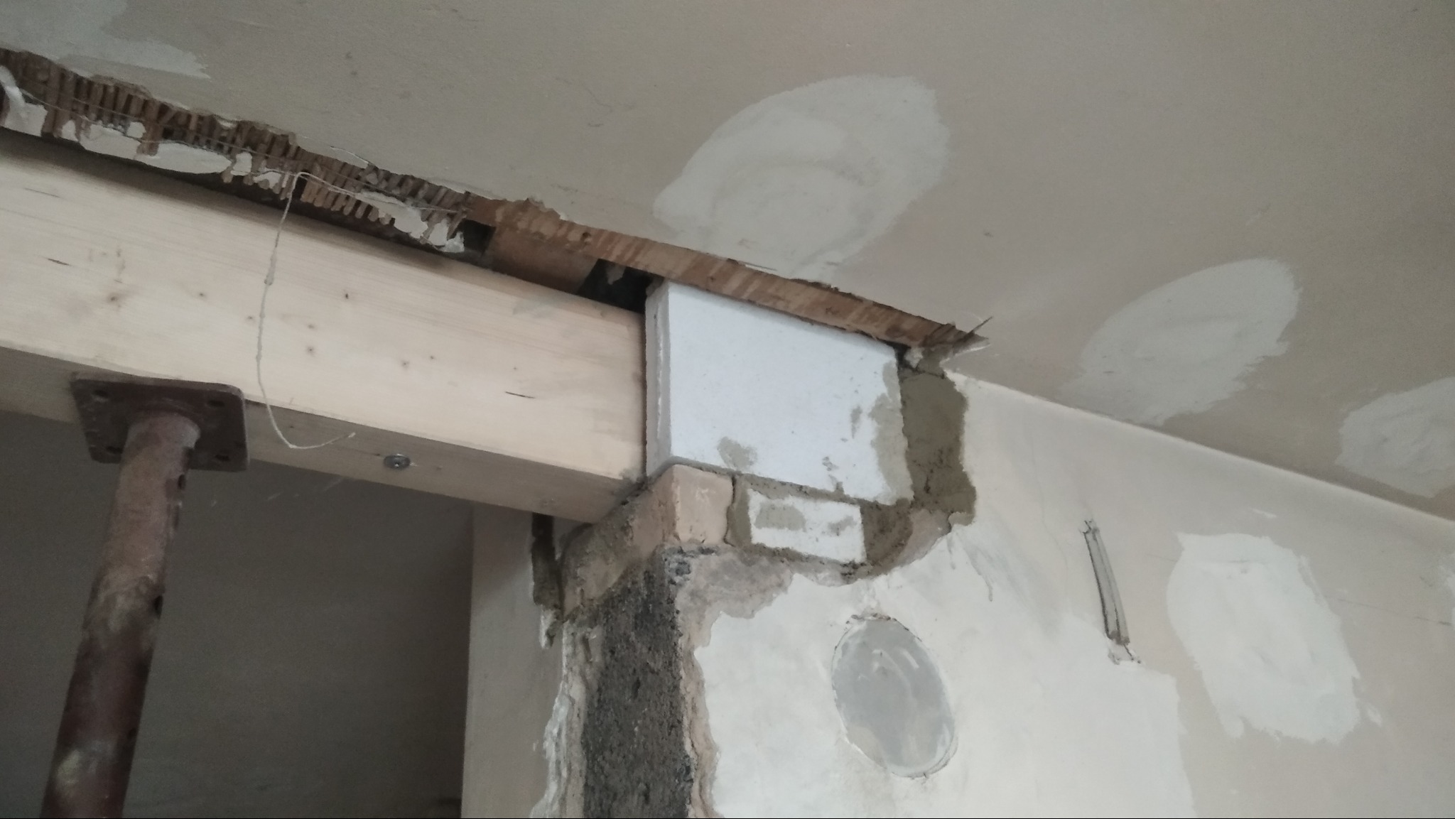 Wzmocnienie otworu w ścianie z użyciem drewnianej belki i podpory. Widoczne fragmenty muru, cegieł i zaprawy. Na suficie ślady po szpachlowaniu.