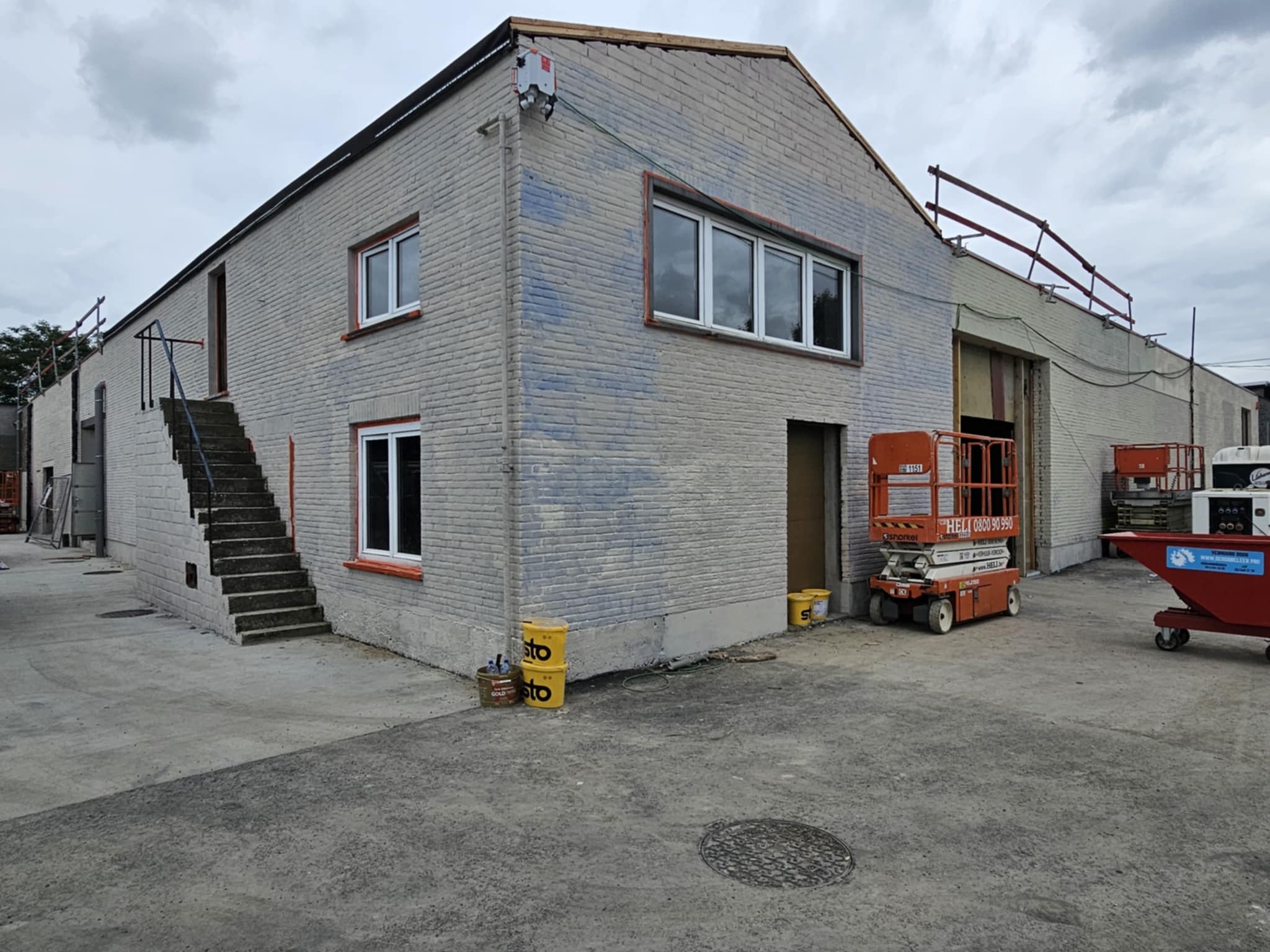 Budynek z cegły w trakcie malowania elewacji na szaro, widoczne ślady po malowaniu, podnośnik koszowy i wiaderka z farbą. Częściowo pomalowana ściana.