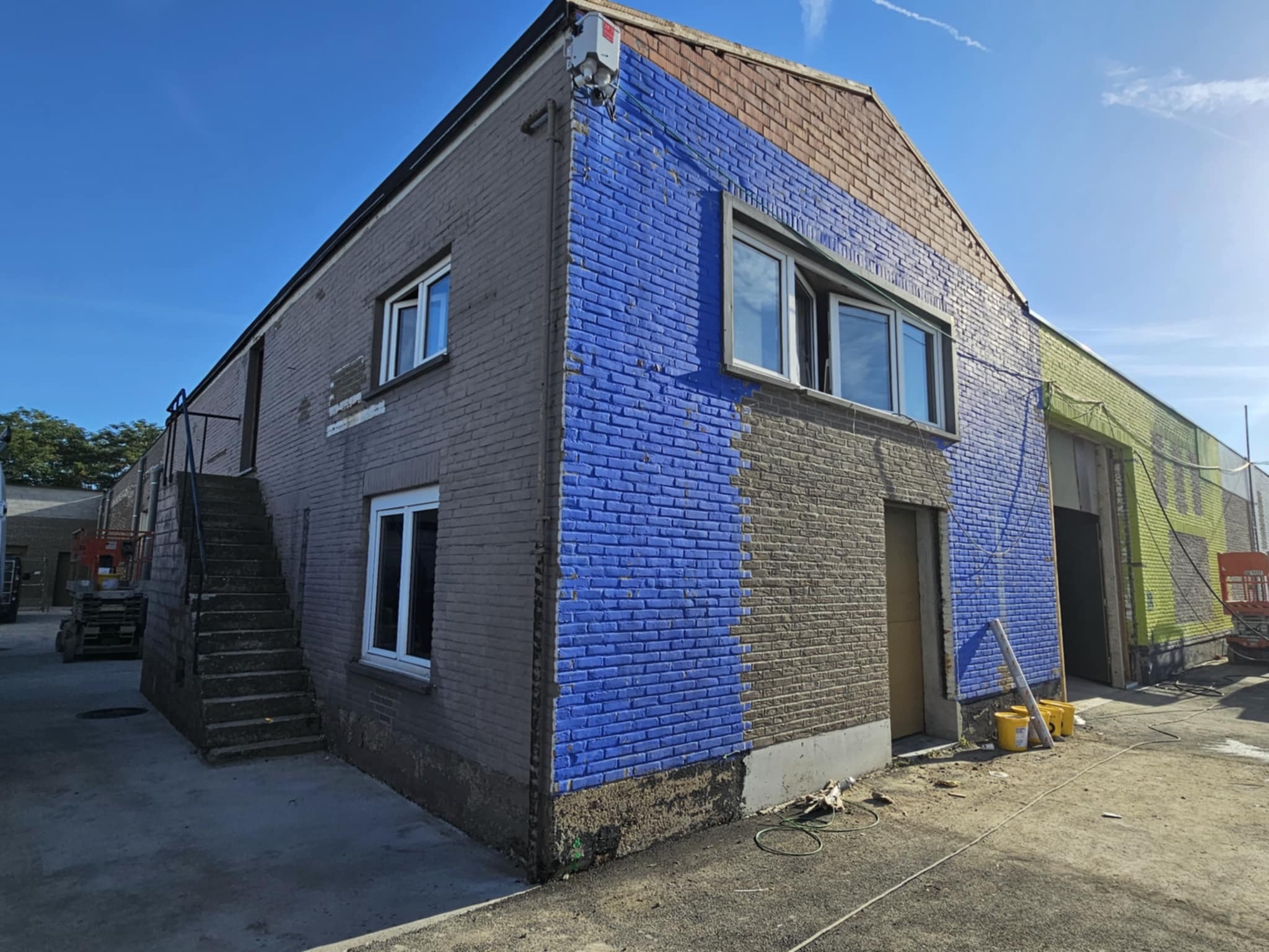 Budynek z fragmentami elewacji w różnych kolorach (szary, niebieski, zielony), ukazujący postęp prac malarskich. Widoczne okna, schody zewnętrzne i sprzęt malarski.