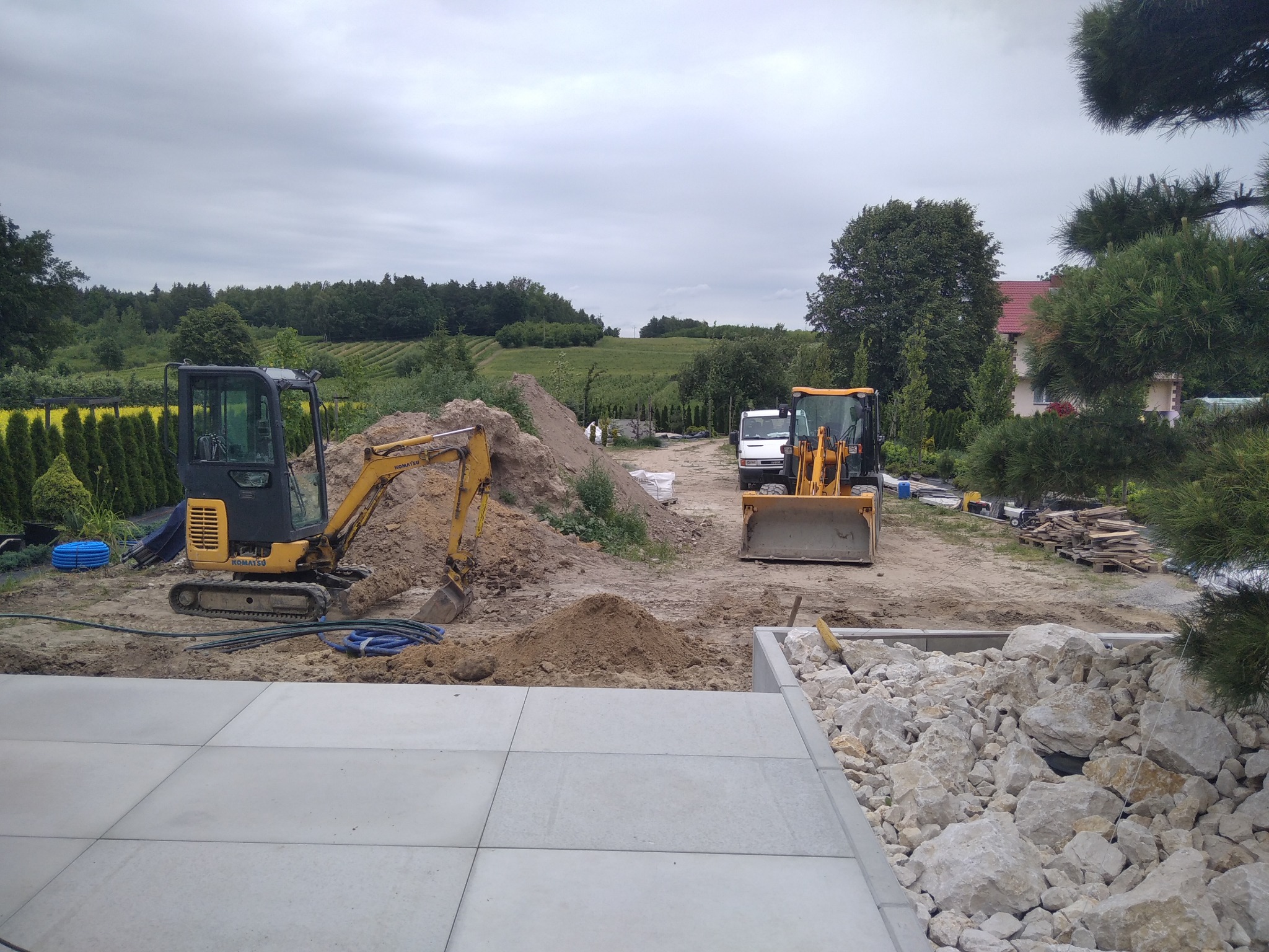 Żółta koparka i ładowarka na placu budowy z ułożonymi płytami chodnikowymi i kamieniami ozdobnymi. W tle zielony krajobraz i zabudowania.