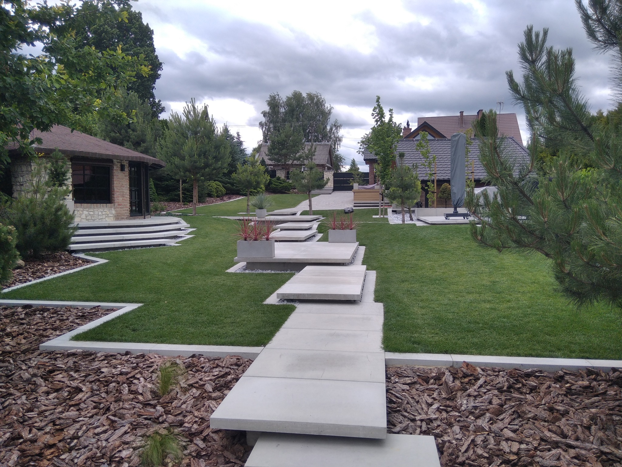 Nowoczesny ogród z trawnikiem, ścieżką z płyt betonowych i rabatami z korą. W tle budynek z kamienia i cegły oraz domy jednorodzinne pod pochmurnym niebem.