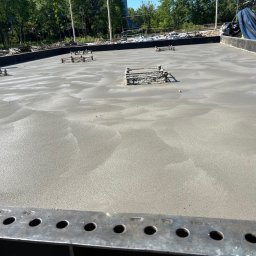 FB Kruczek - Świeżo wylany fundament z widocznymi elementami zbrojenia, przygotowany do dalszych prac budowlanych. Widok z bliska, z metalową listwą w dolnej części kadru.