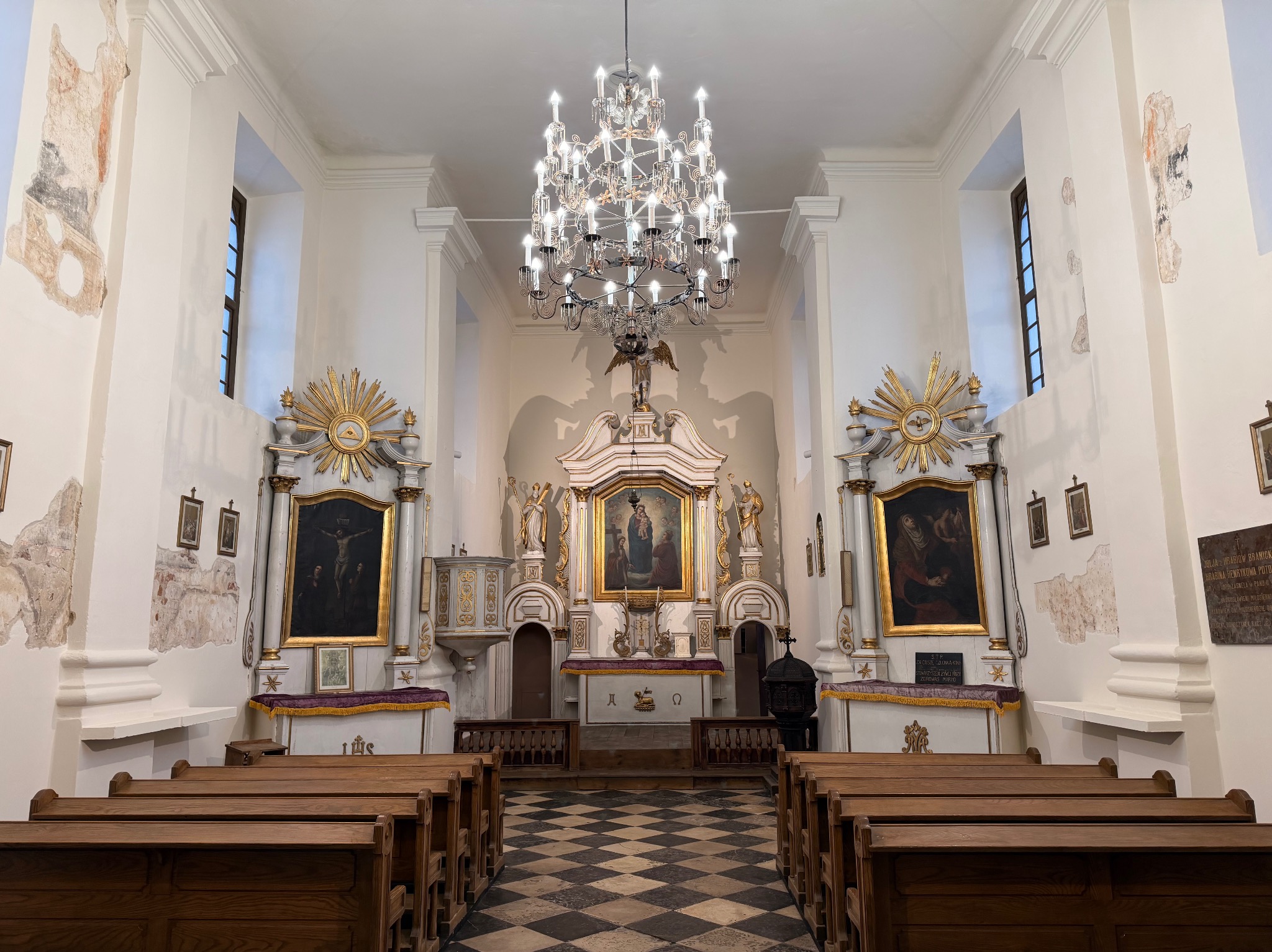 Wnętrze kościoła z drewnianymi ławkami, ołtarzem i żyrandolem. Widoczne obrazy, dekoracje i szachownica na podłodze. Ściany z widocznymi śladami renowacji.