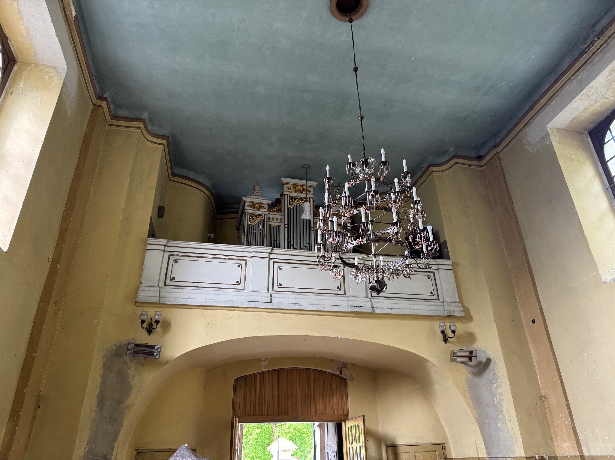 Wnętrze kościoła w trakcie malowania: ściany w odcieniach żółci, sufit błękitny, widoczne organy i żyrandol. Ślady po malowaniu na ścianach.