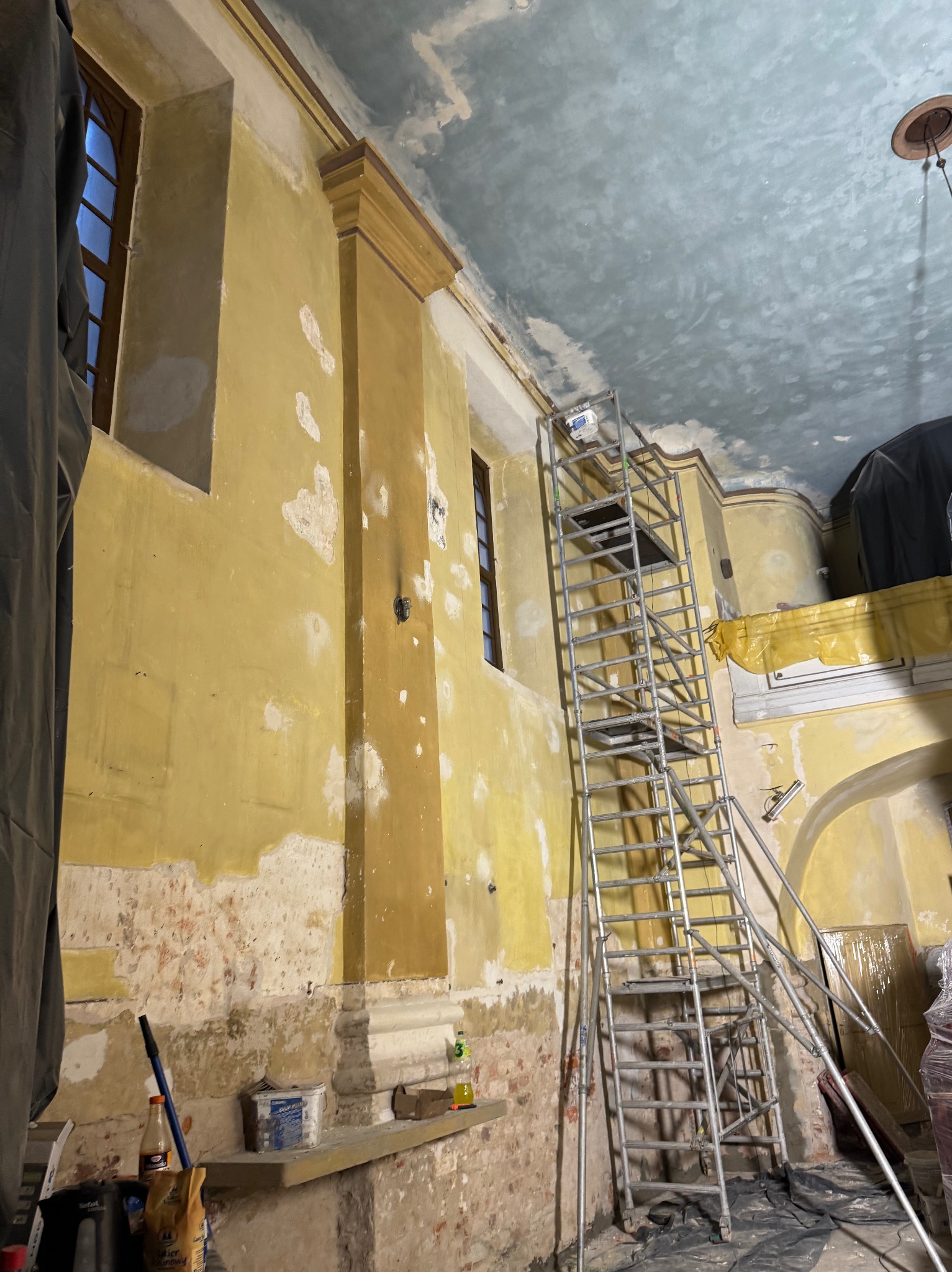 Wnętrze w trakcie renowacji: żółte ściany z ubytkami farby, rusztowanie aluminiowe, fragment okna i sufit z widocznymi śladami prac malarskich.