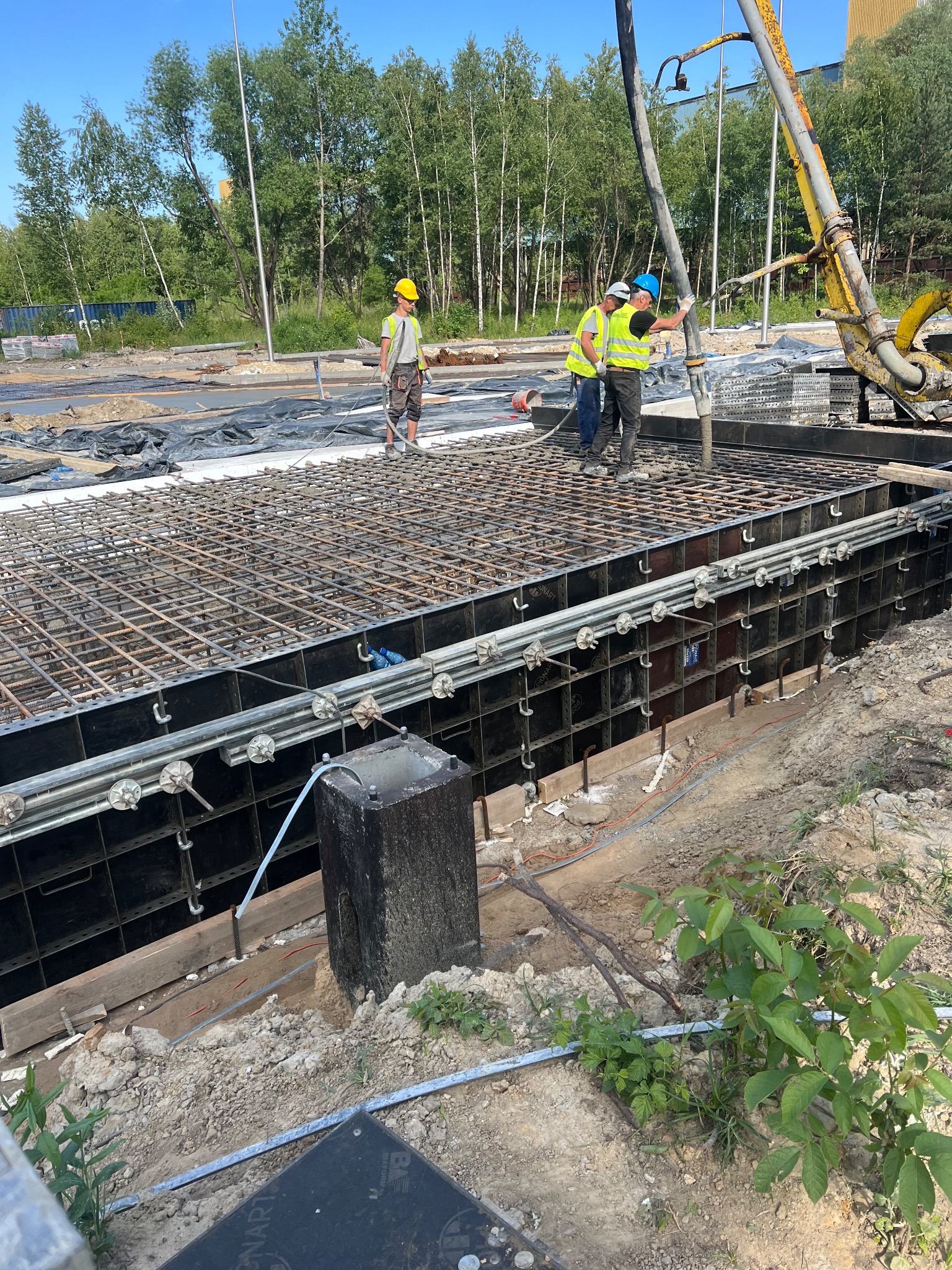 Wylewanie betonu do szalunku zbrojonego na budowie. Trzech robotników w kaskach i kamizelkach nadzoruje proces. W tle drzewa i niebo.