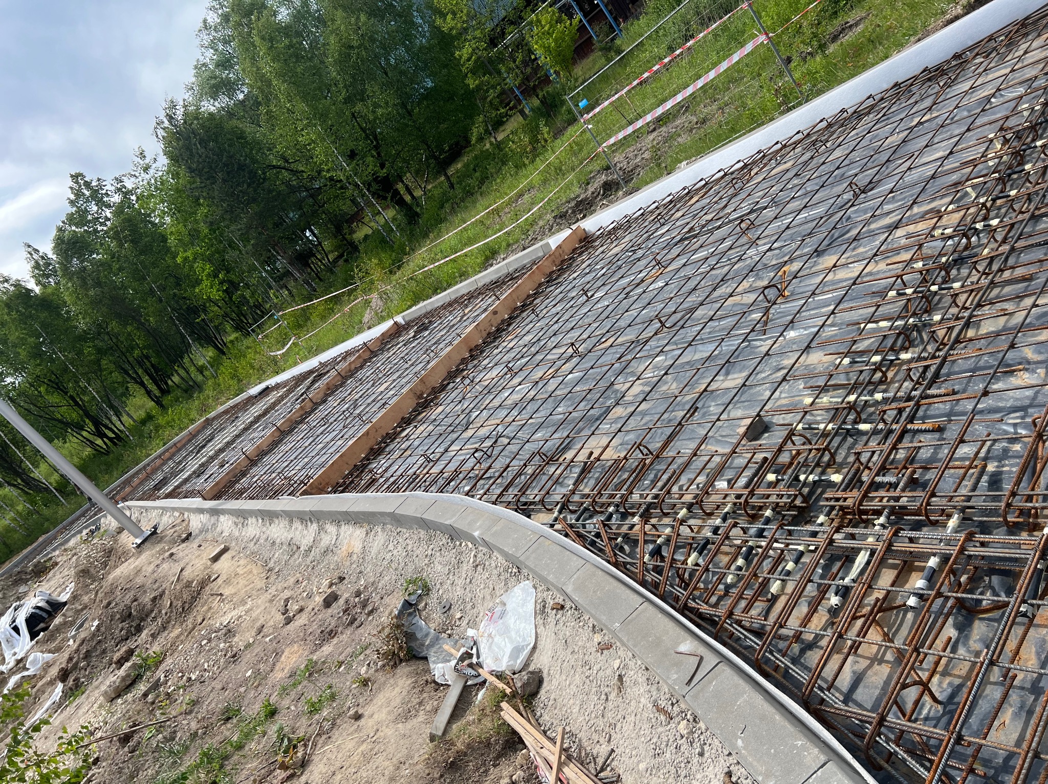 Zbrojenie fundamentu z ułożoną siatką stalową, widok z góry na przygotowany teren budowy z zielenią w tle. Elementy szalunku drewnianego i krawężnik betonowy.