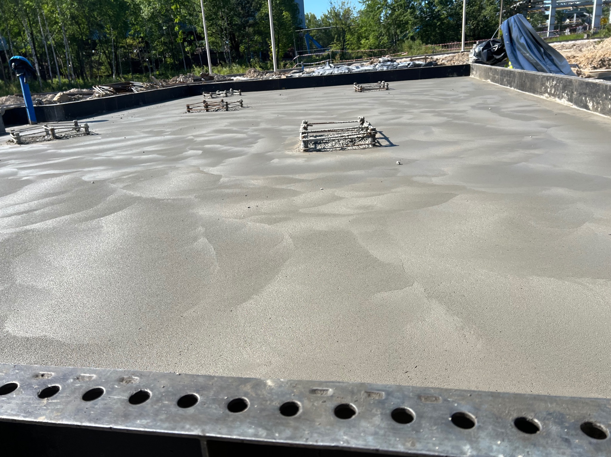 Świeżo wylany fundament z widocznymi elementami zbrojenia, przygotowany do dalszych prac budowlanych. Widok z bliska, z metalową listwą w dolnej części kadru.