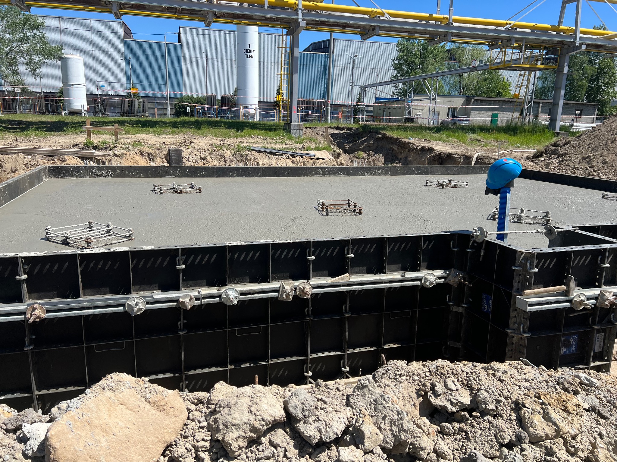Zalewanie betonem fundamentów w szalunku systemowym, widoczne zbrojenie i niebieski kask na rurze. W tle budynki przemysłowe i instalacje rurowe.