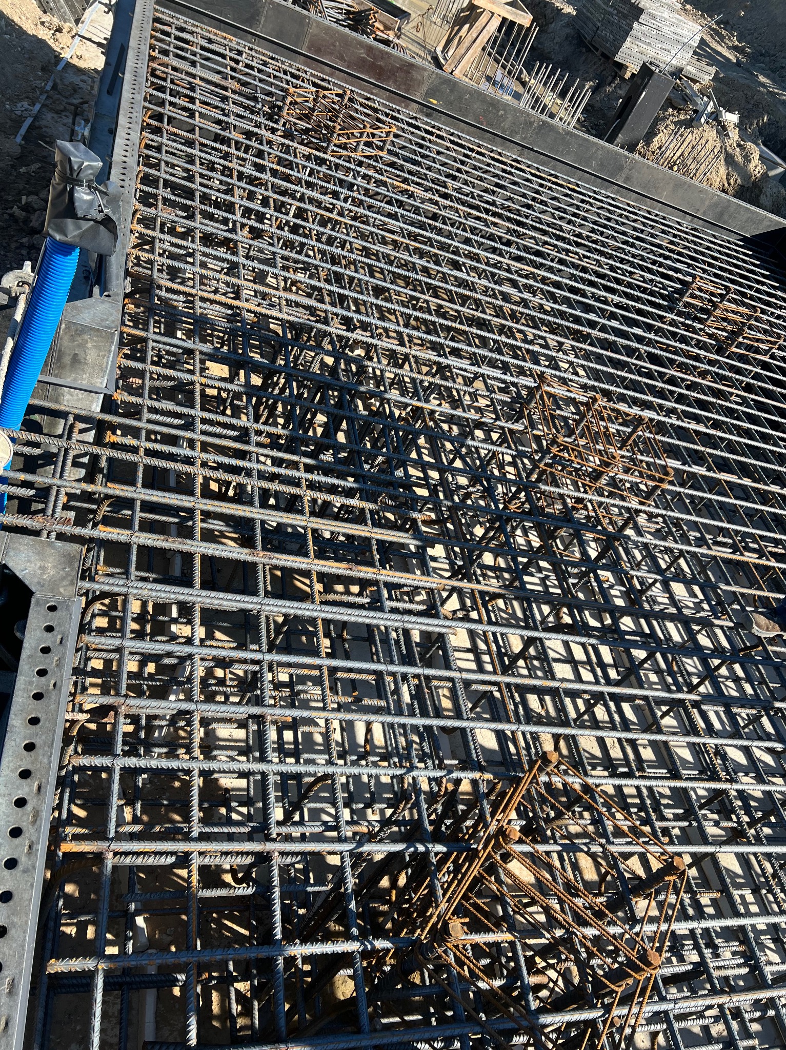 Siatka zbrojeniowa ułożona w szalunku fundamentu, widok z góry. Elementy konstrukcyjne i niebieska rura w tle. Detal zbrojenia.