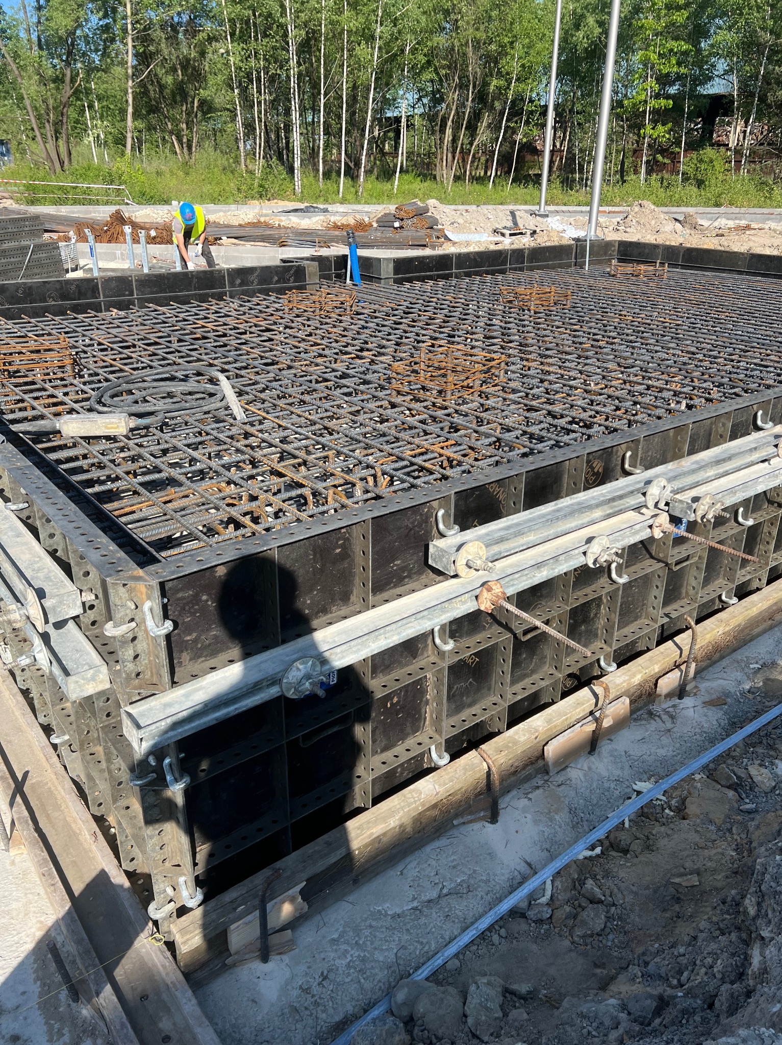 Zbrojenie fundamentu w szalunku systemowym, widok z góry. Na drugim planie pracownik w kasku i kamizelce odblaskowej. W tle drzewa.