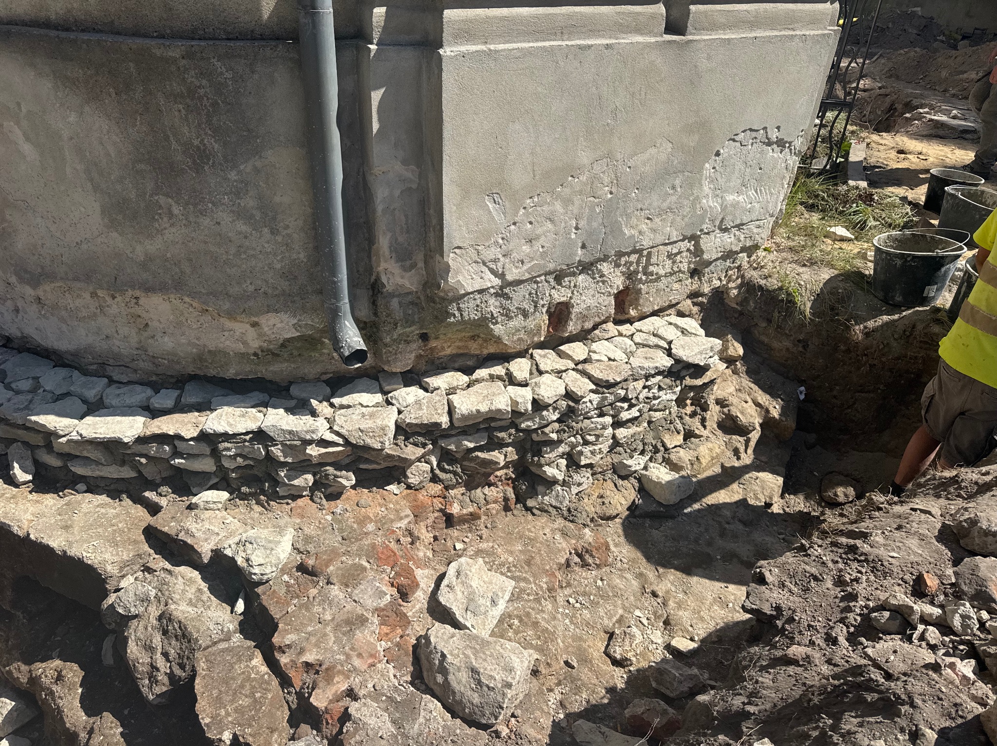 Odkopany fundament starego budynku z widocznymi warstwami kamienia i cegły, fragment rynny spustowej, ziemia i gruz wokół wykopu. Prace ziemne przy fundamencie.