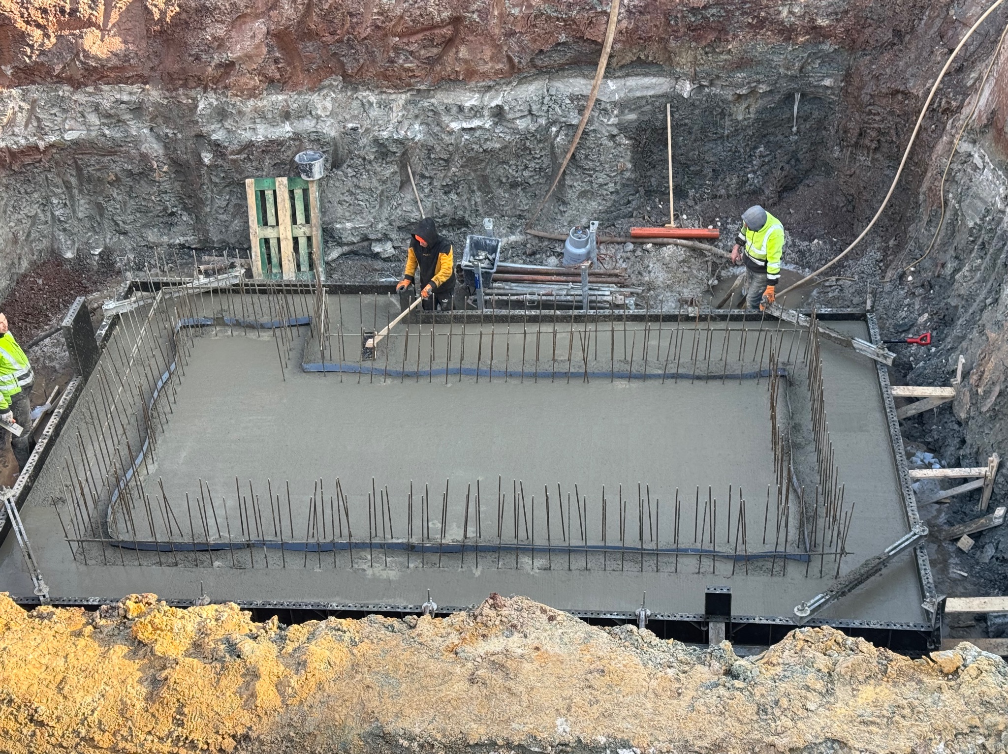 Wylany betonowy fundament z wystającymi prętami zbrojeniowymi, widok z góry na wykop, dwóch pracowników w odblaskowych ubraniach wyrównuje powierzchnię betonu.