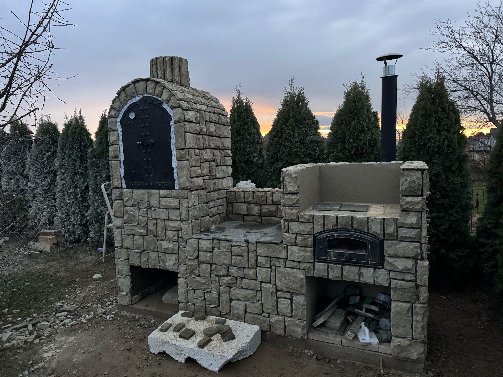 Kamienny piec ogrodowy z czarnymi drzwiczkami i kominem, otoczony tujami, w trakcie budowy. Widoczne narzędzia i materiały budowlane.