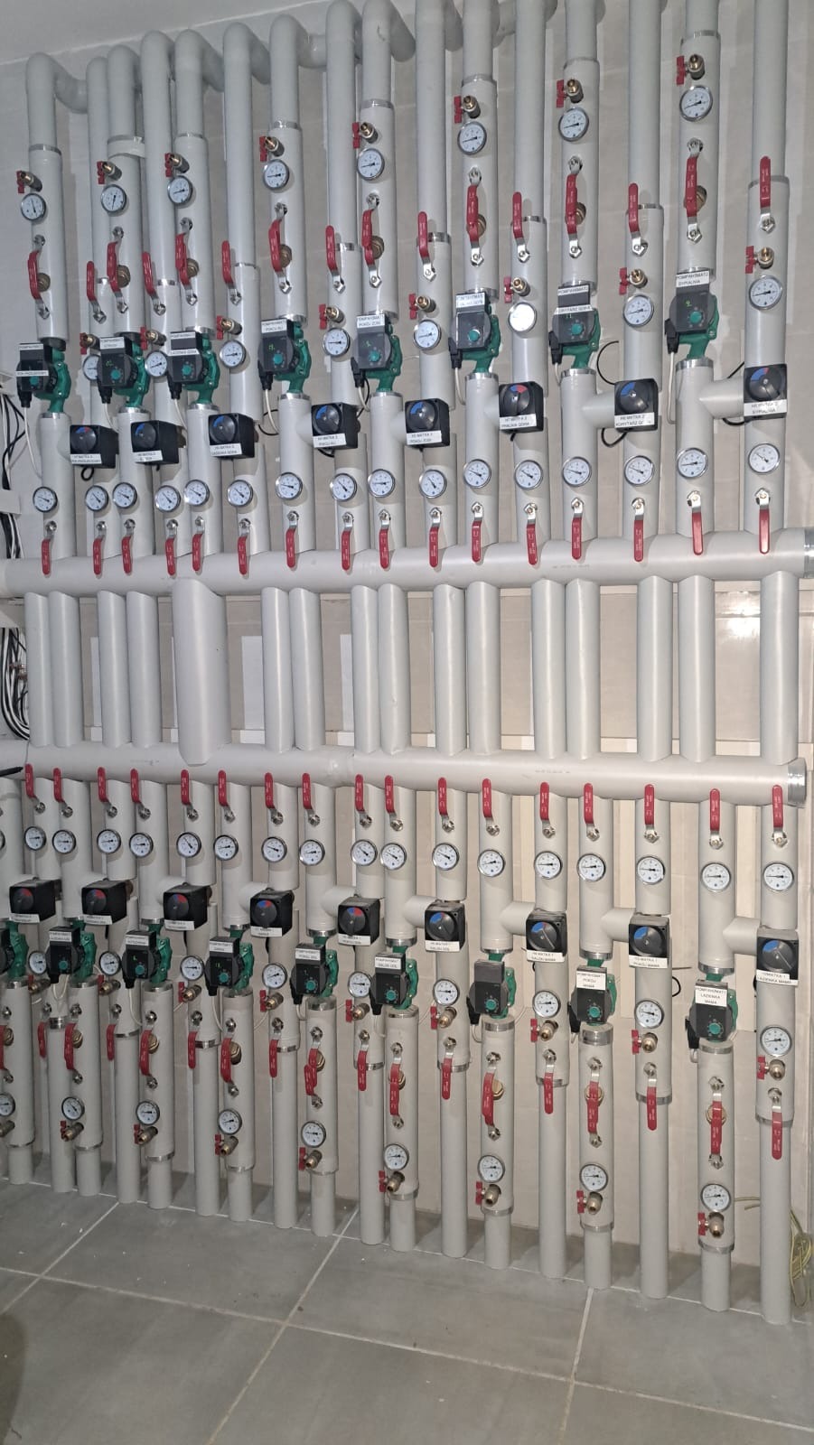 Rozbudowana instalacja grzewcza z szarych rur, zaworów i manometrów, tworząca symetryczny układ na ścianie. Widoczne pompy obiegowe i liczne punkty kontrolne.