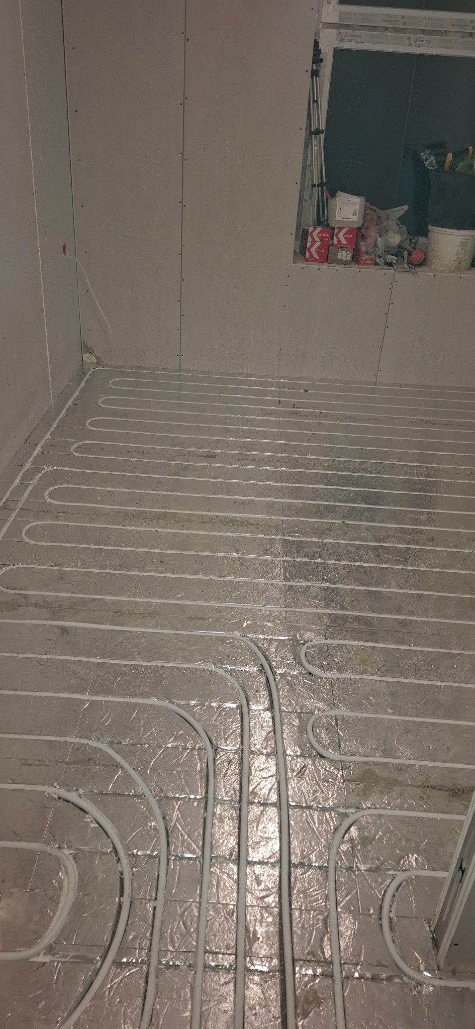 Białe rury ogrzewania podłogowego ułożone na srebrnej folii izolacyjnej w surowym wnętrzu z płyt kartonowo-gipsowych, przed wylaniem posadzki. Widok z góry.