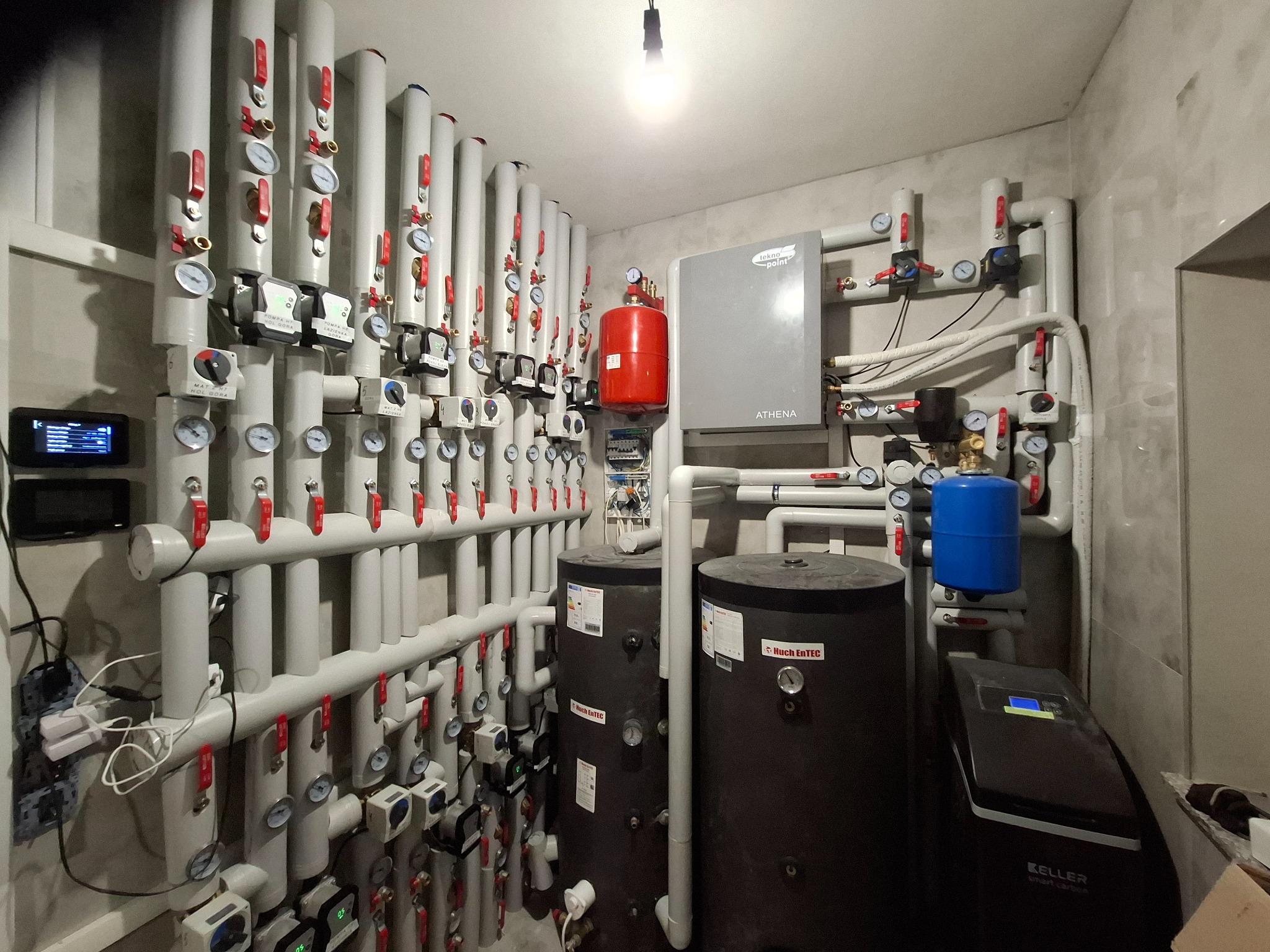 Złożona instalacja grzewcza w kotłowni: rzędy rur z zaworami, dwa czarne zbiorniki, szary piec Athena, niebieski zbiornik i czarny zmiękczacz Keller Smart Carbon.
