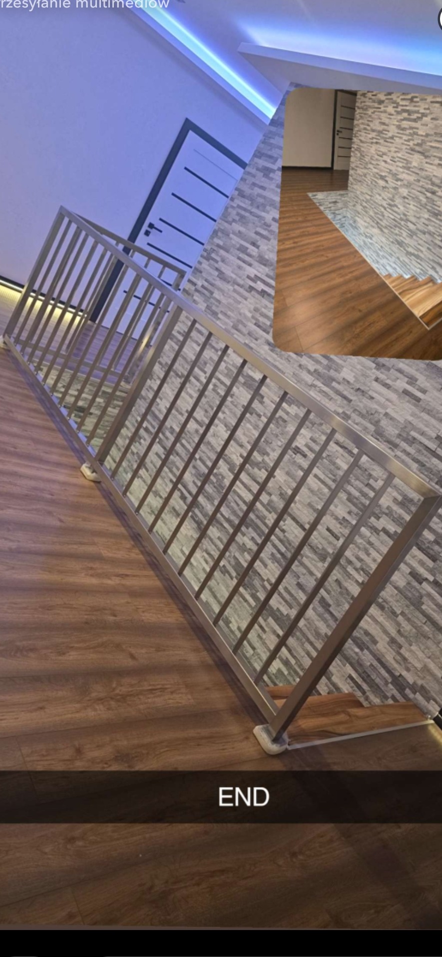 Nowoczesna, metalowa balustrada schodowa z pionowymi szczeblami, zamontowana wewnątrz budynku. Widoczne drewniane schody i ściana wyłożona kamieniem dekoracyjnym.