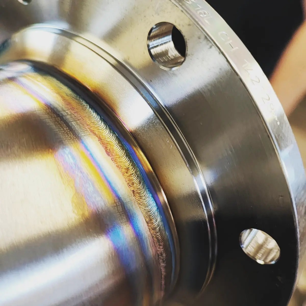 Błyszczące, metalowe złącze rurowe z widocznym, precyzyjnym spoiwem spawalniczym, opalizującym kolorami tęczy. Widoczne otwory montażowe i napisy techniczne.