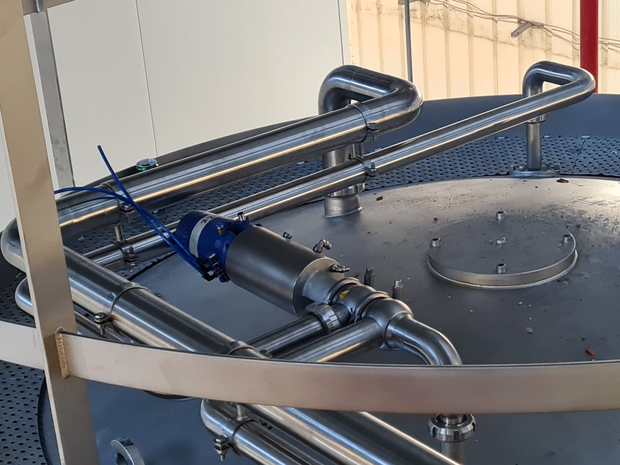 Błyszczące, metalowe rury i elementy instalacji przemysłowej na tle zbiornika. Widoczne złącza, mocowania i silnik elektryczny. Ujęcie z góry, oświetlenie naturalne.