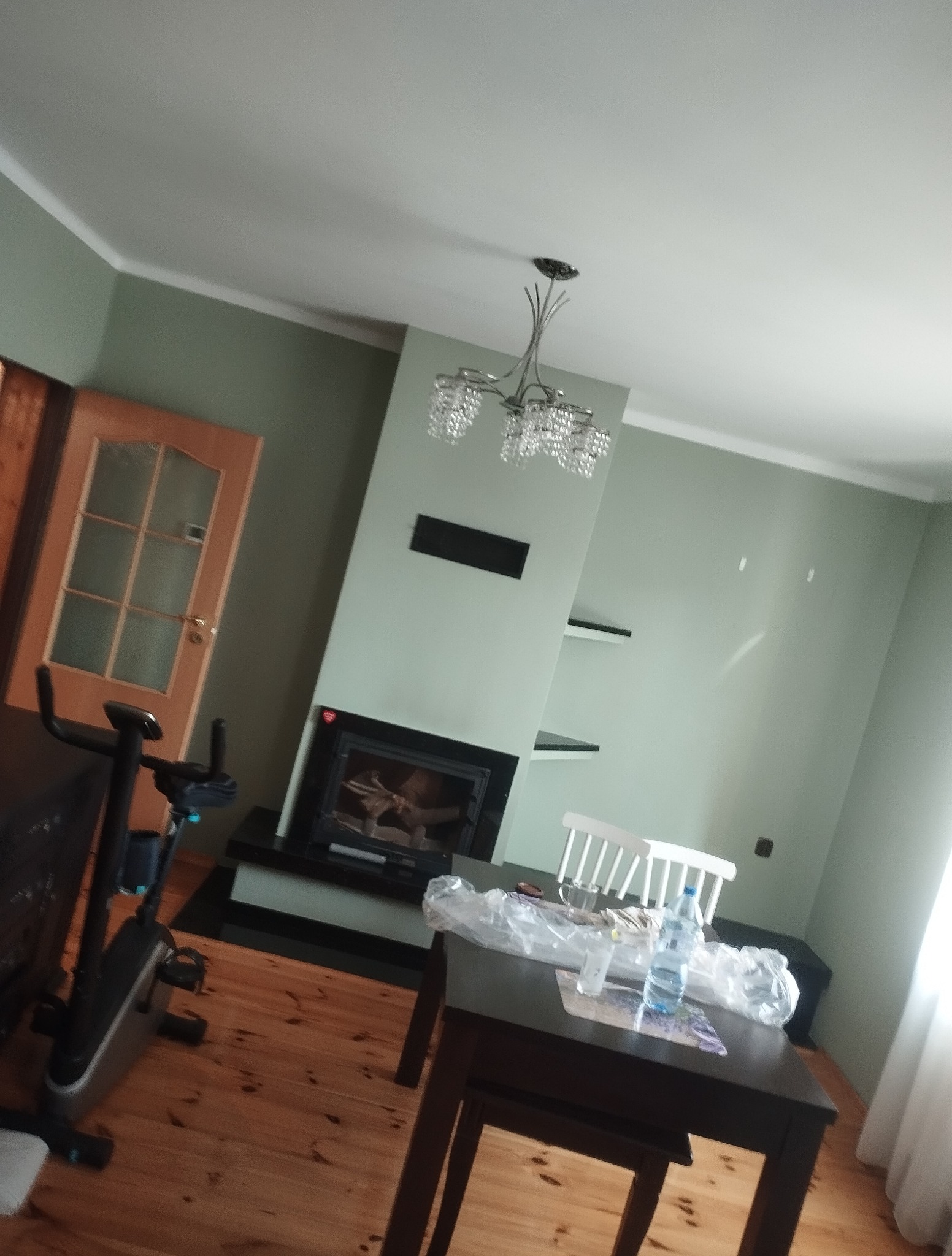 Jasne wnętrze z kominkiem i drewnianą podłogą. Nad stołem kryształowy żyrandol. Ściany w odcieniach zieleni. Widoczny rower treningowy.