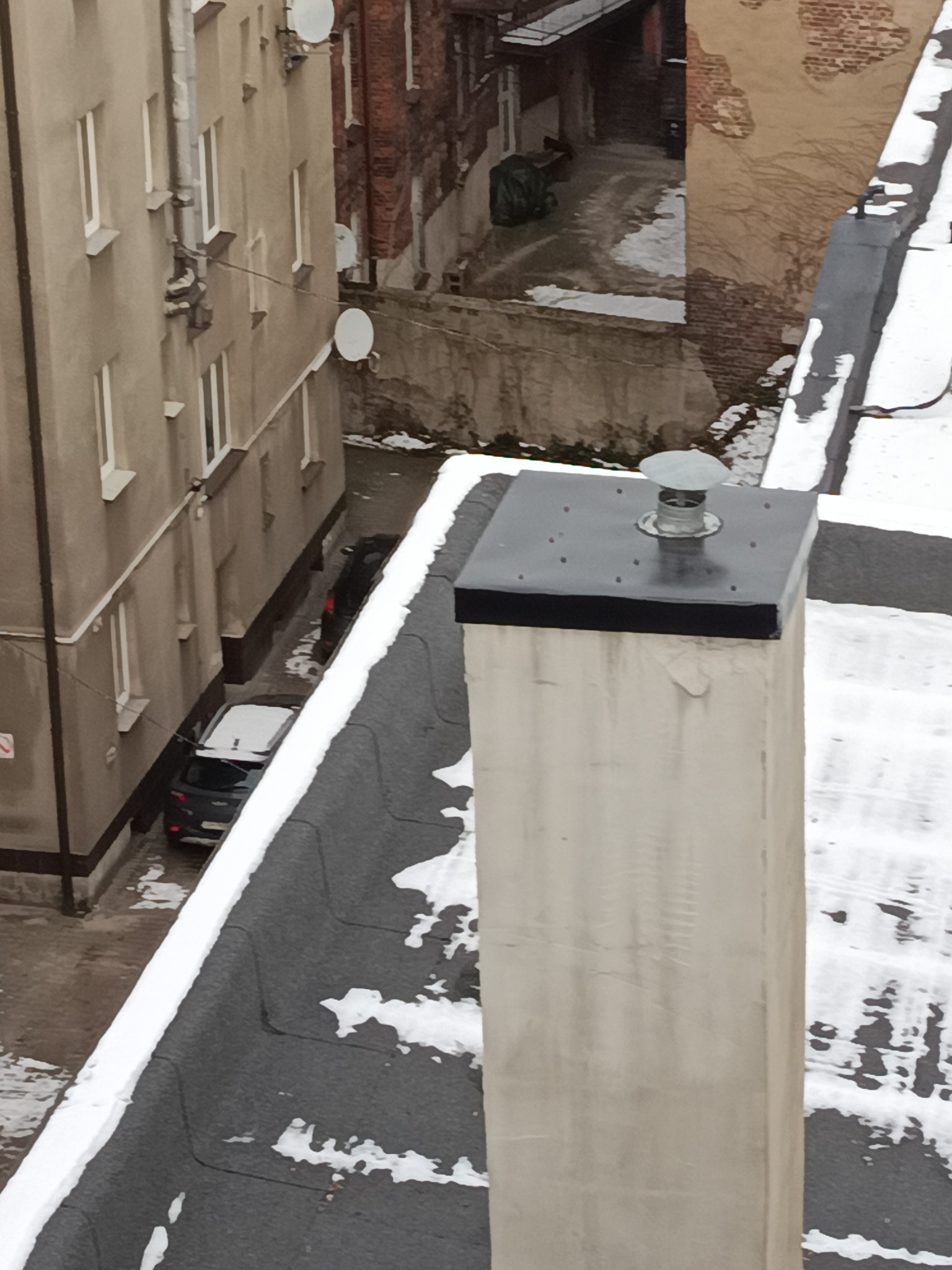 Widok z góry na płaski dach pokryty papą z kominem wentylacyjnym, częściowo pokryty śniegiem. W tle budynki mieszkalne i podwórko z zaparkowanymi samochodami.