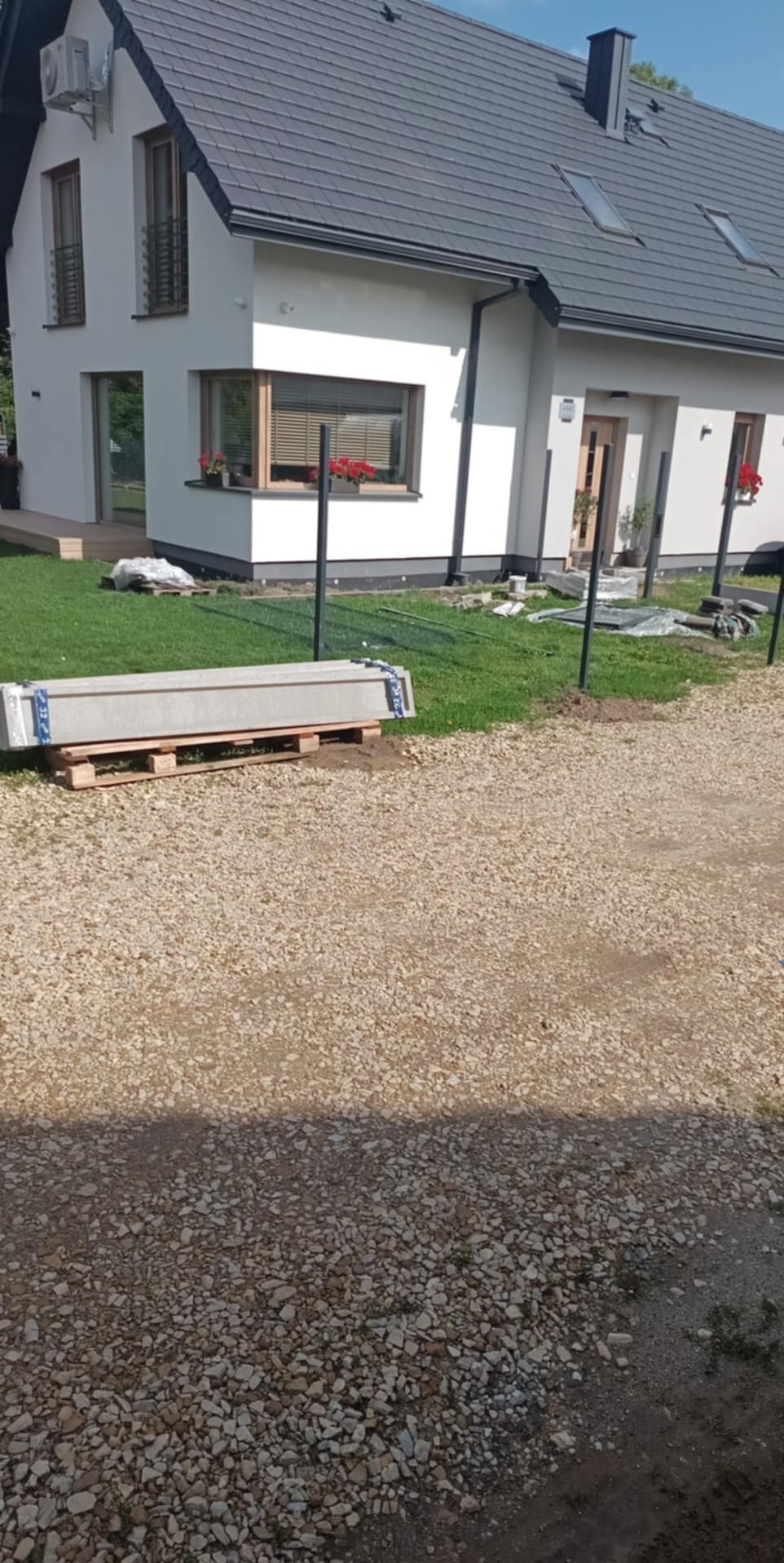 Montaż słupków ogrodzeniowych na posesji. Widoczne słupki w ziemi i paleta z elementami ogrodzenia na tle nowoczesnego domu z szarą dachówką.