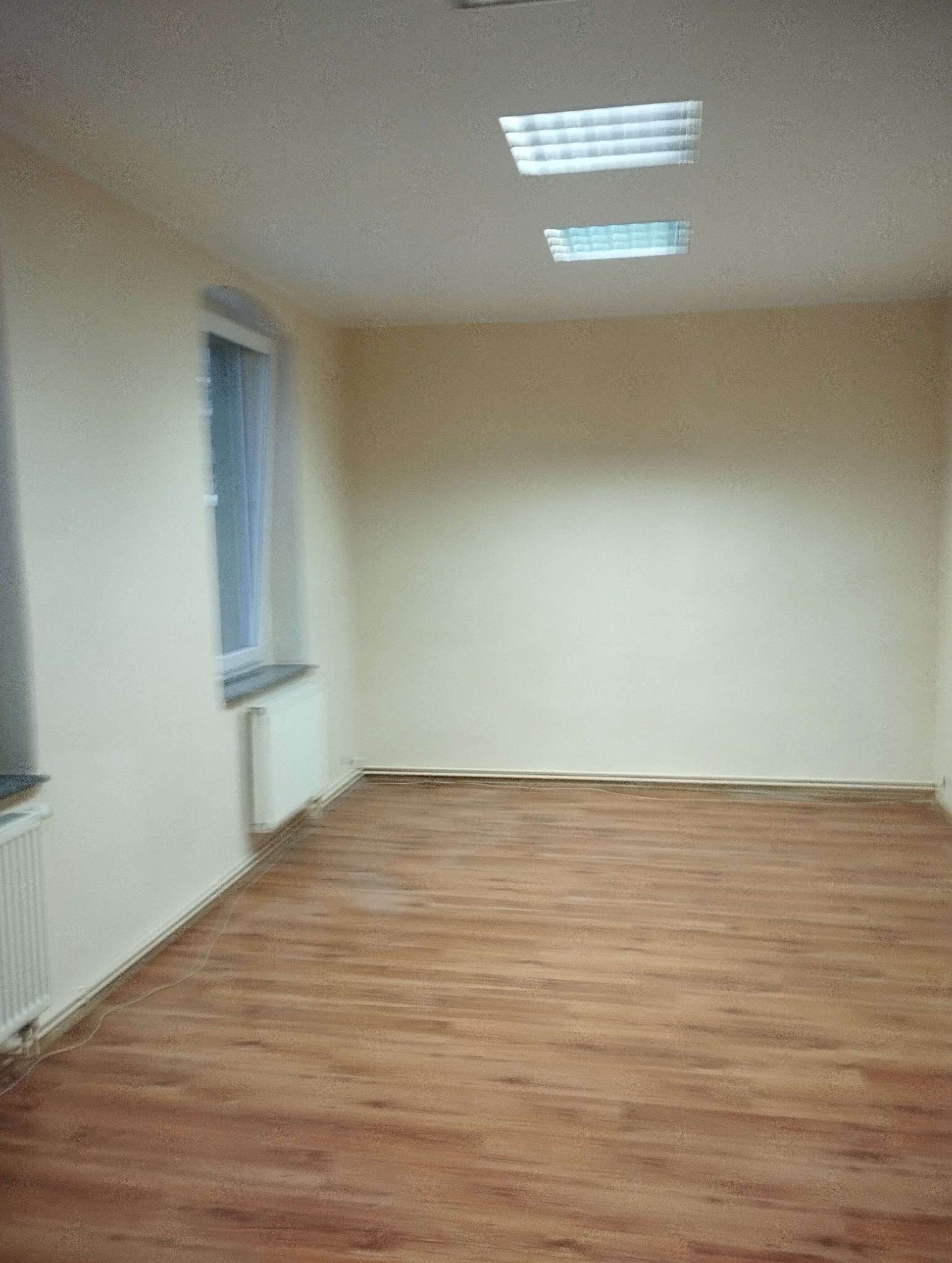 Puste, jasne wnętrze z panelami podłogowymi, grzejnikiem pod oknem i dwoma lampami sufitowymi. Ściany w odcieniach beżu. Widok na puste pomieszczenie przed wykończeniem.