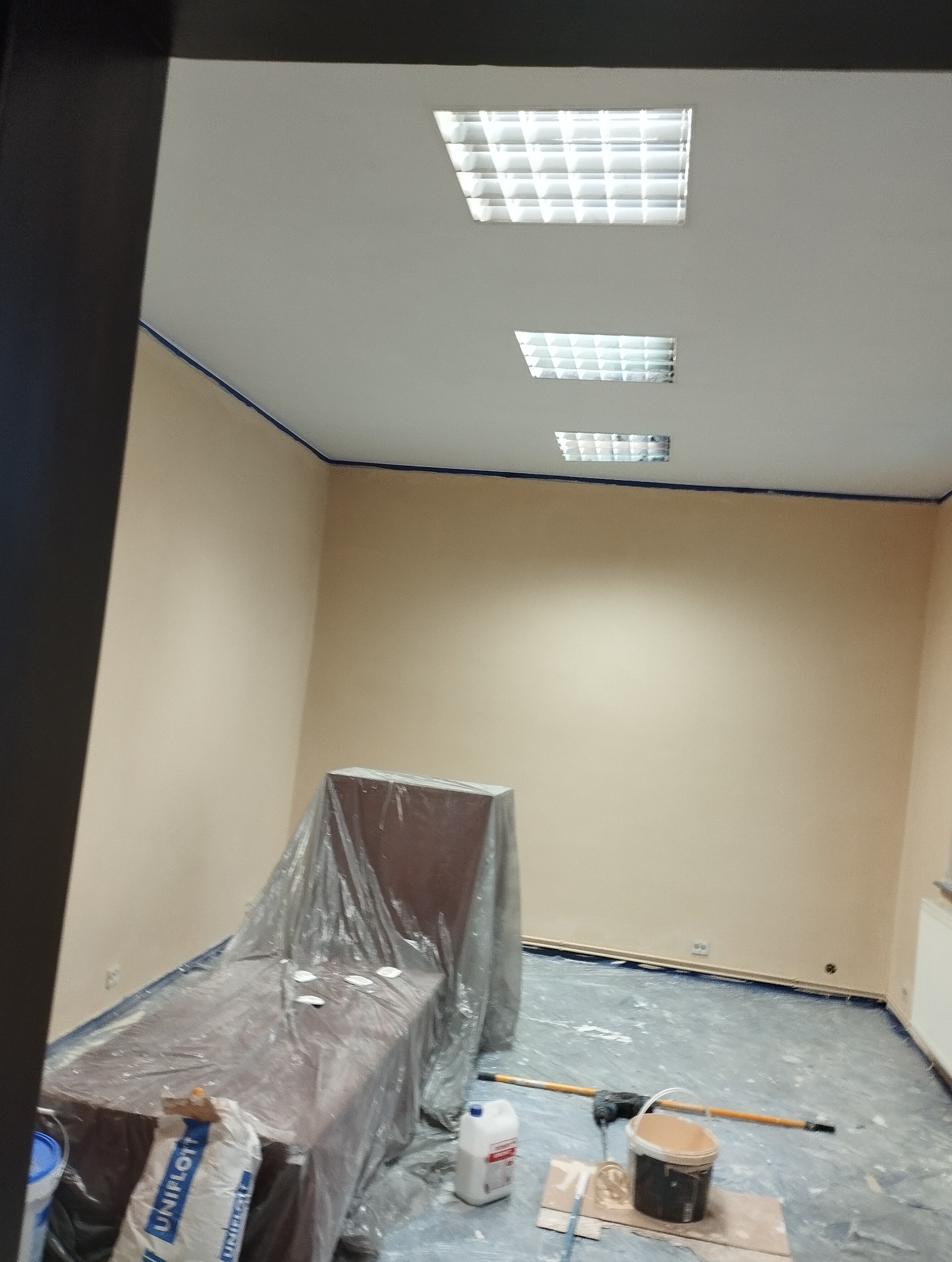 Wnętrze po malowaniu ścian na beżowo, zabezpieczone meble folią, widoczne narzędzia malarskie i taśma malarska przy suficie. Oświetlenie sufitowe w kształcie kwadratów.