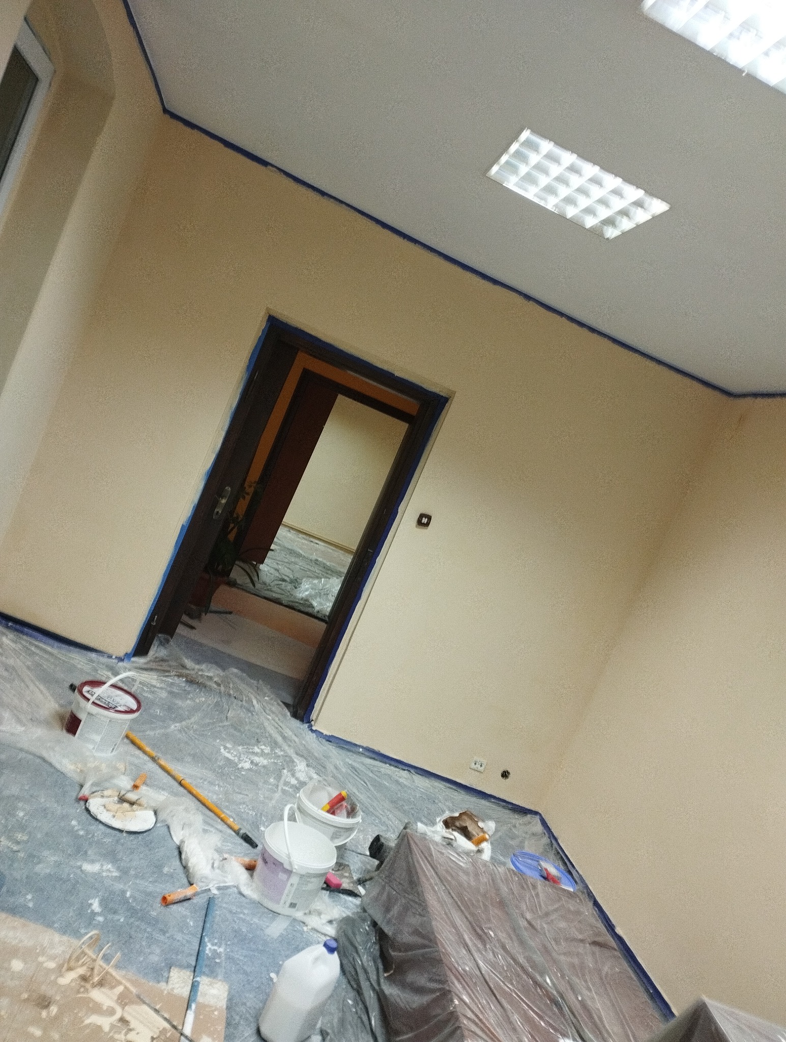 Pokój w trakcie malowania: ściany w beżowym odcieniu, zabezpieczone folią meble, widoczne narzędzia malarskie i puszki z farbą. Niebieska taśma malarska na krawędziach.