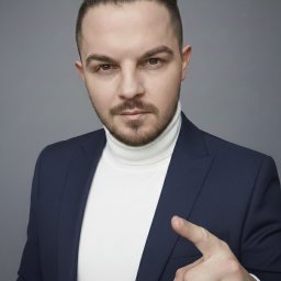 Paweł Kwiatek - Portret mężczyzny w granatowej marynarce i białym golfie, wskazującego palcem w stronę widza. Na palcu złoty pierścień. Gładkie tło, profesjonalne oświetlenie.