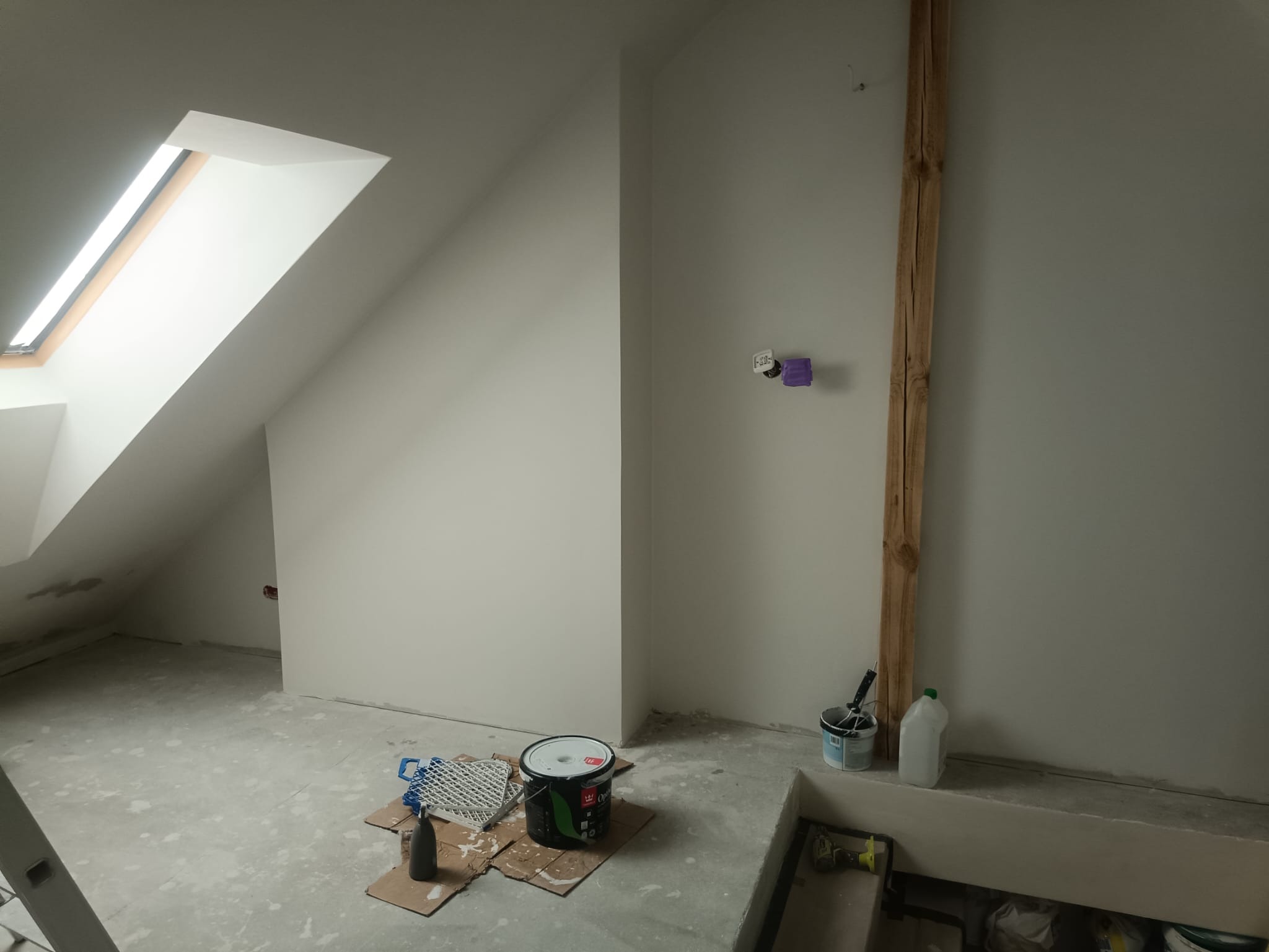 Poddasze po malowaniu na biało, widoczne okno dachowe, drewniana listwa, puszka elektryczna, wiadro z farbą i akcesoria malarskie. Świeżo pomalowane ściany.