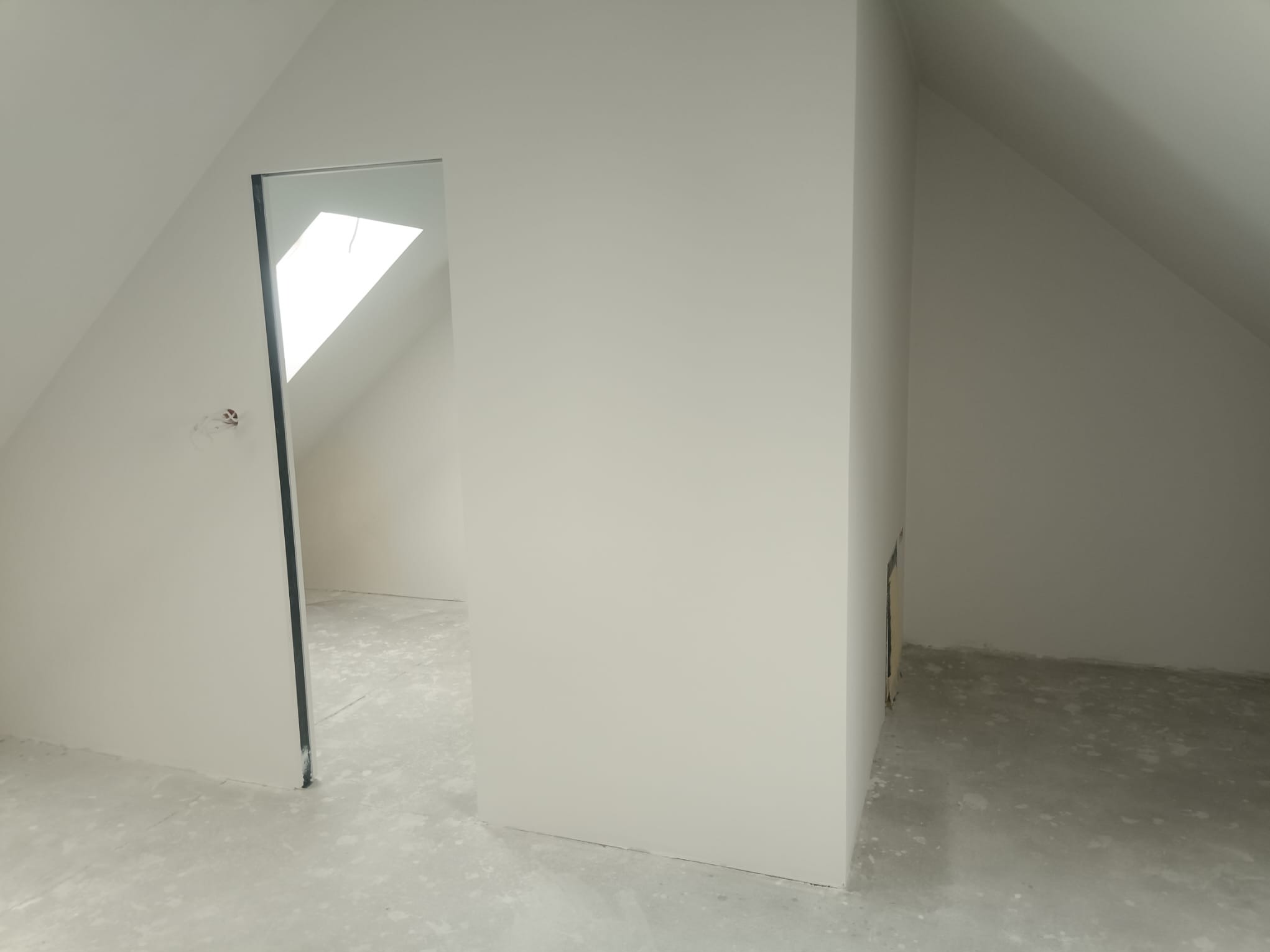 Wnętrze poddasza po malowaniu na biało, widoczne ściany, podłoga i ościeżnica drzwiowa. Minimalistyczny styl, jasne oświetlenie, surowe wykończenie.