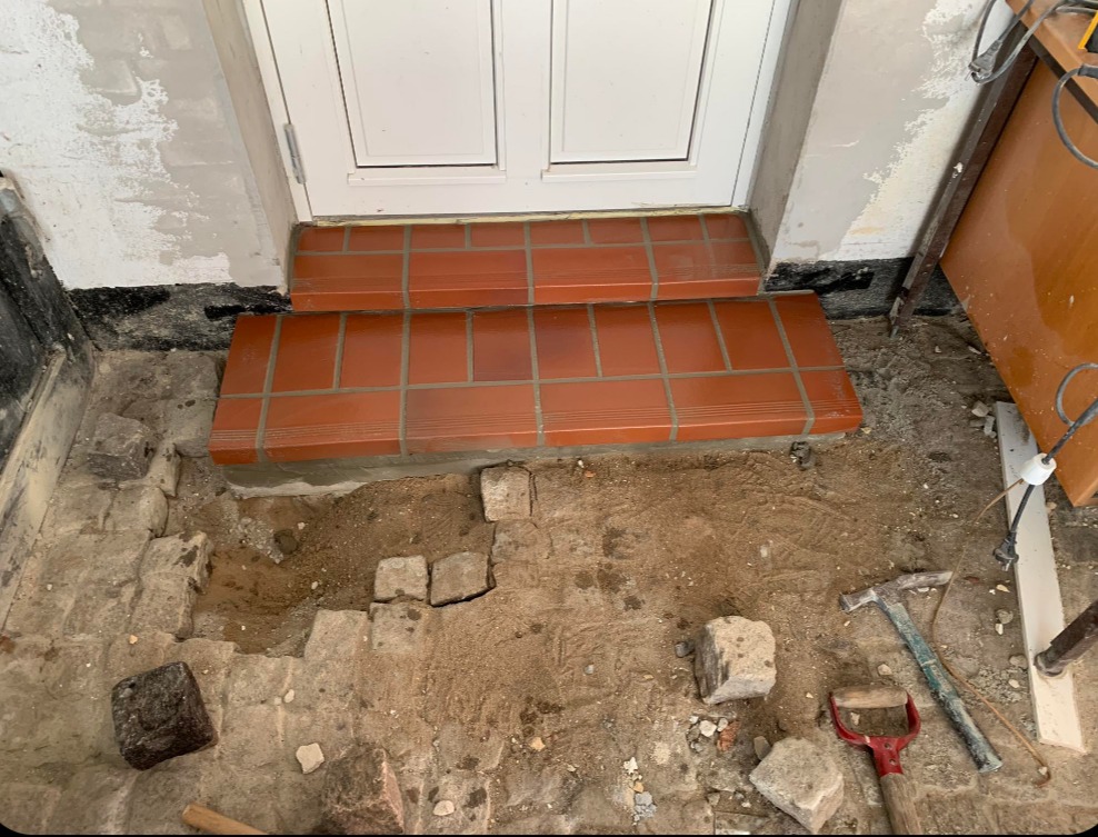 Ułożone płytki schodowe w odcieniach terakoty przy białych drzwiach wejściowych, widoczne narzędzia i gruz, etap prac wykończeniowych.