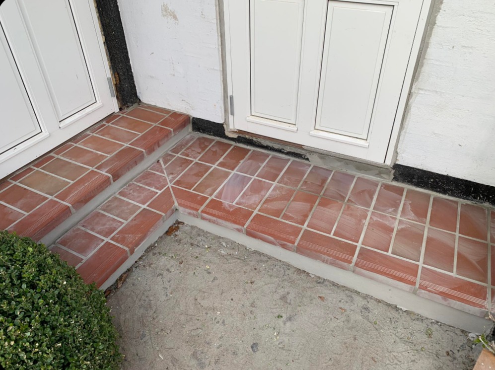 Wykończone płytkami schody wejściowe z terakoty, prowadzące do białych drzwi. Widoczne fugi i fragment zieleni po lewej stronie. Ujęcie z góry, blisko detalu.