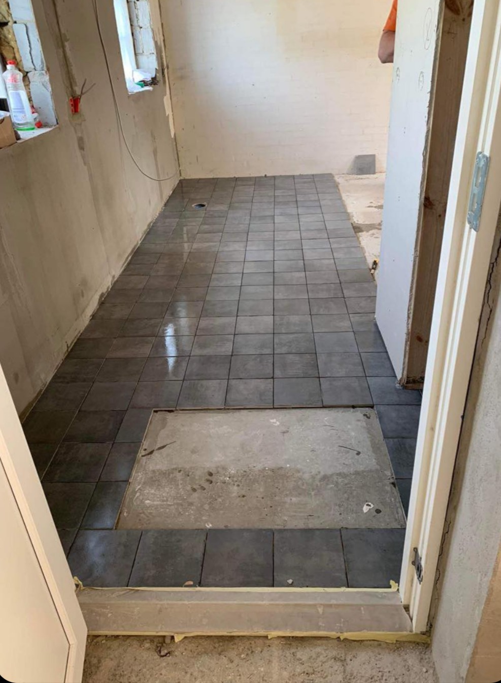 Układanie ciemnych, kwadratowych płytek na podłodze w pomieszczeniu w trakcie remontu. Widoczny otwór rewizyjny bez płytek i fragment ściany w surowym stanie.